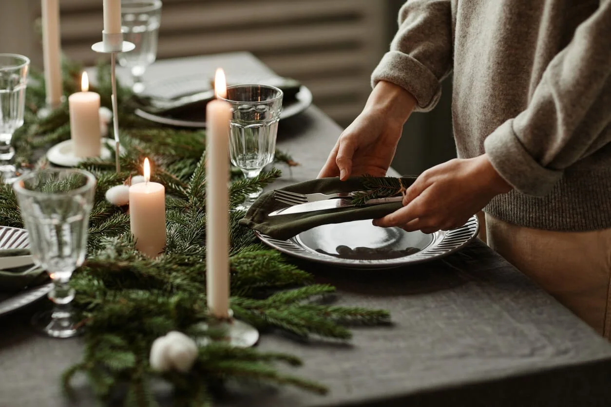 A person in a gray knit sweater folds a napkin onto a plate on a holiday table set with candles, glasses, and evergreen branches