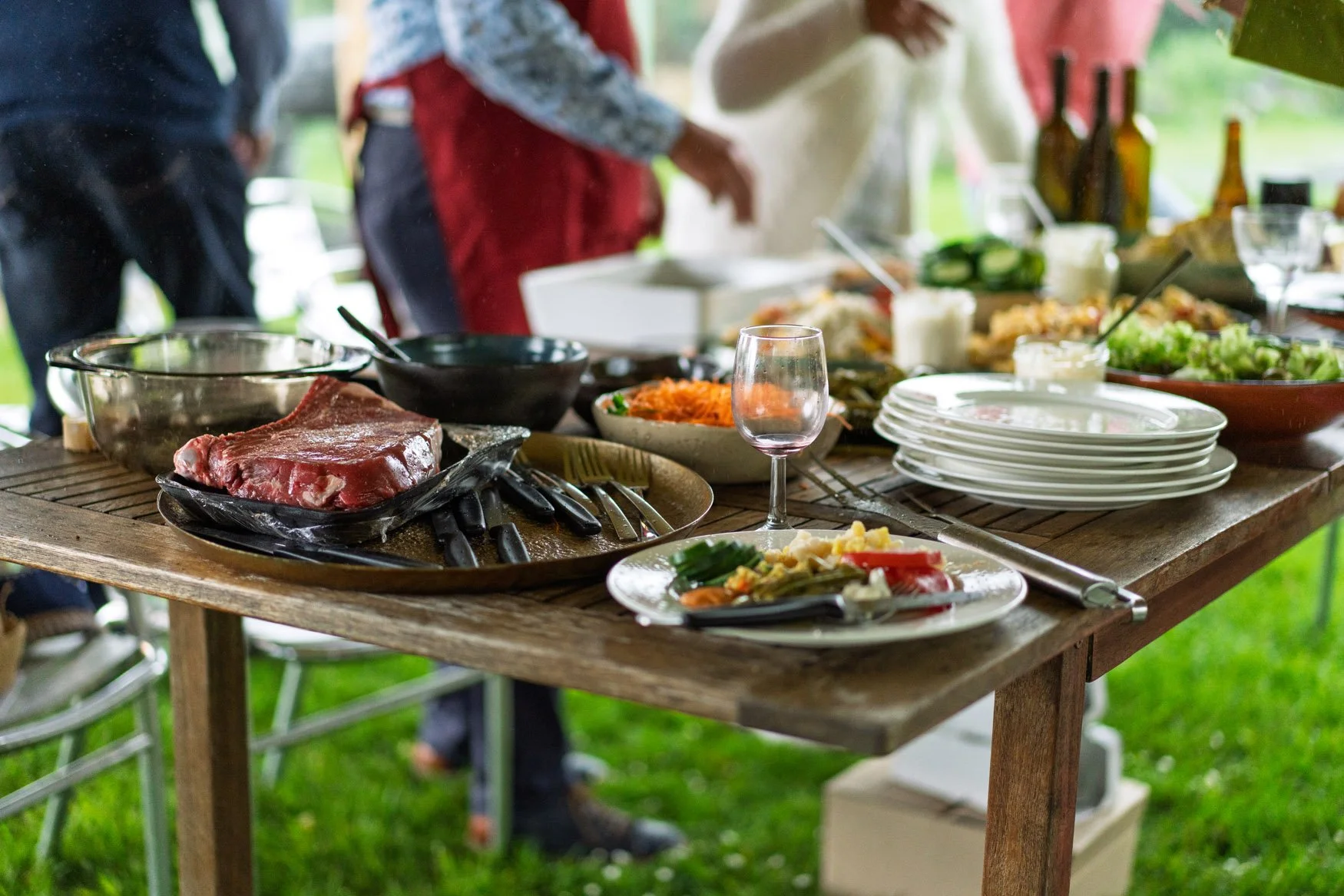 A rustic wooden table set for a barbecue meal outdoors, with raw meat, vegetables, plates, glasses, and various side dishes, and people in the background.