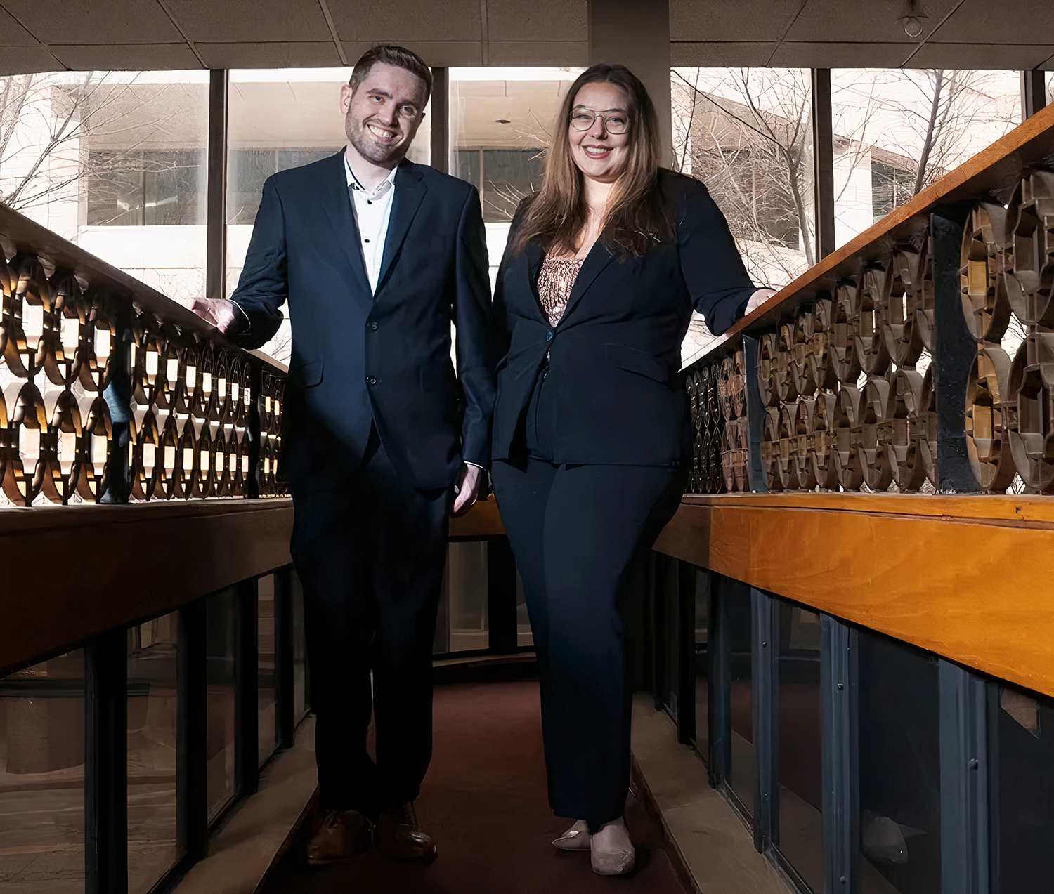 A man and woman dressed in formal business attire pose together on a balcony with decorative wooden railing. They are smiling, standing close to each other with the man's left arm resting on the railing. Behind them are large windows with trees visible outside.