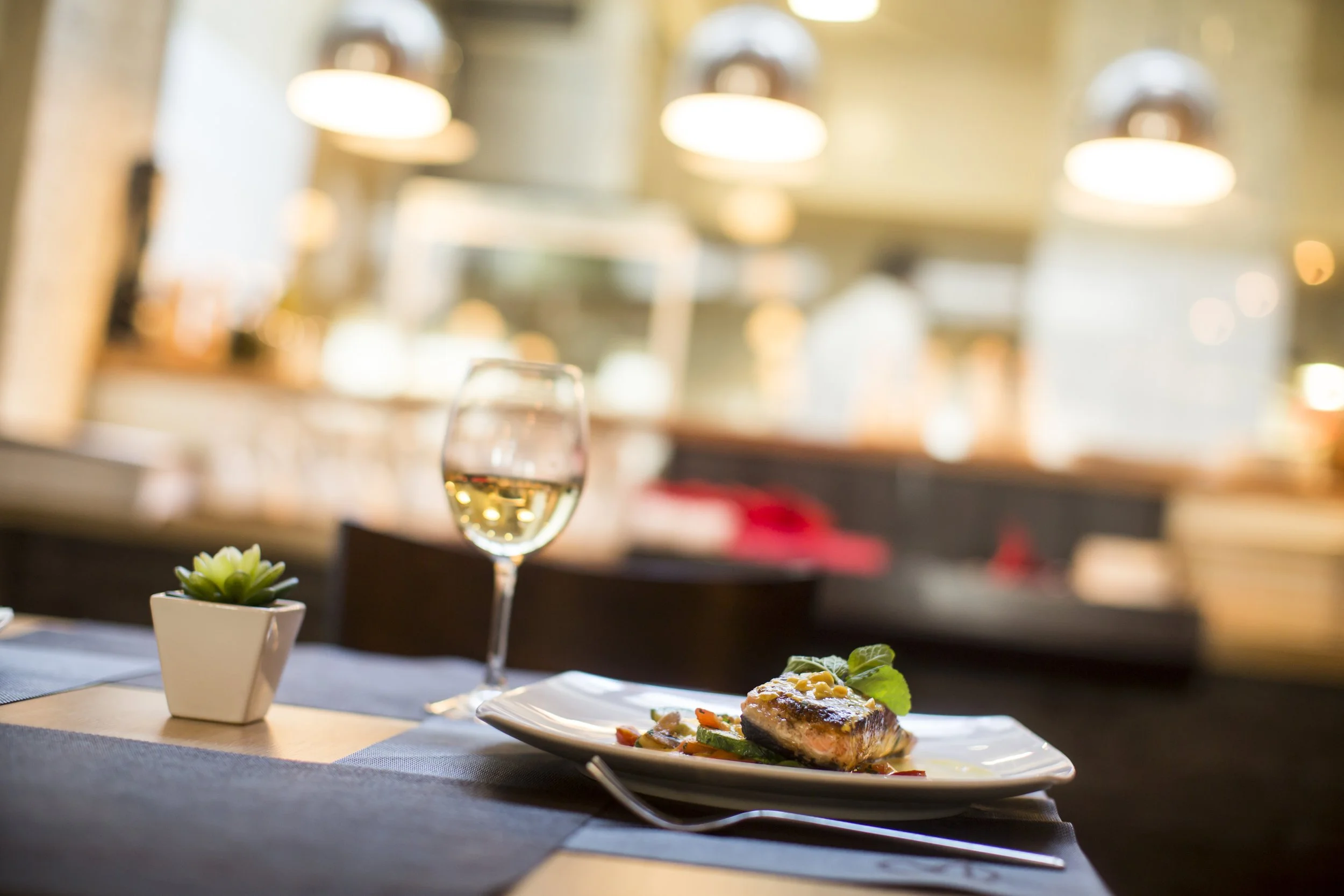 A dining table in a restaurant with a glass of white wine, a small potted succulent plant, and a plate of fish with vegetables. The background features warm lighting and blurred interior details.