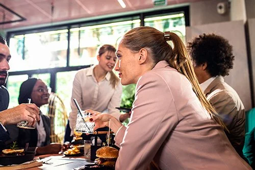 Group of five people sitting and standing at a table in a restaurant or cafe, engaged in conversation, with food and drinks on the table and large windows in the background.