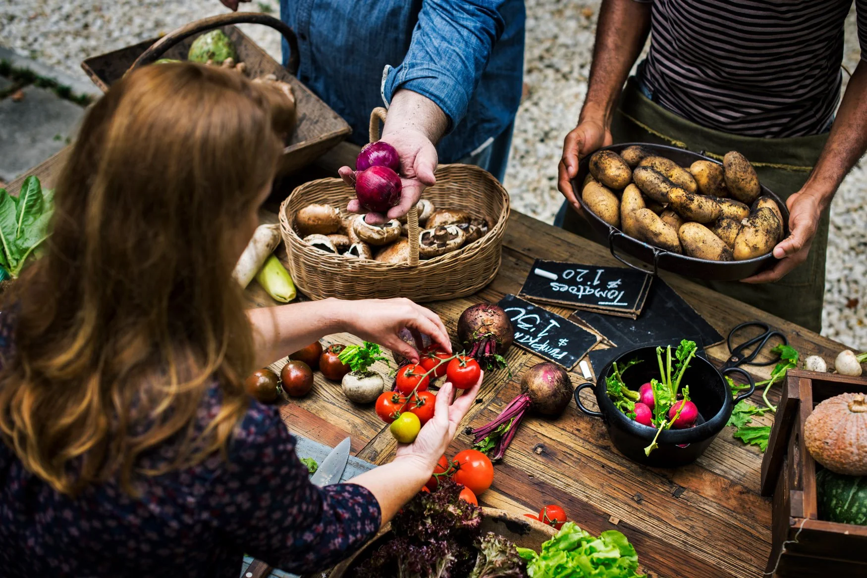 People at a farmers market interacting with fresh vegetables including tomatoes, radishes, potatoes, and onions on a wooden table.