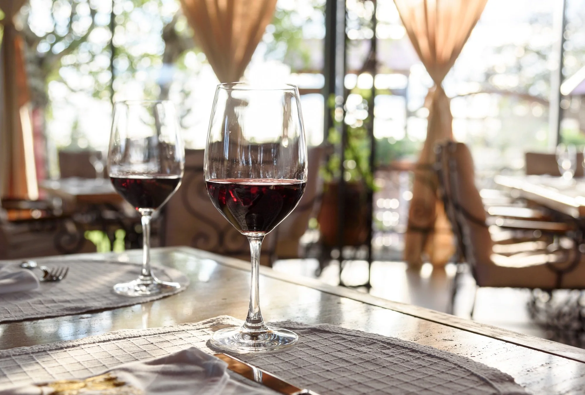 Two glasses of red wine on a table in a cozy restaurant with wooden furniture and large windows showing trees outside.