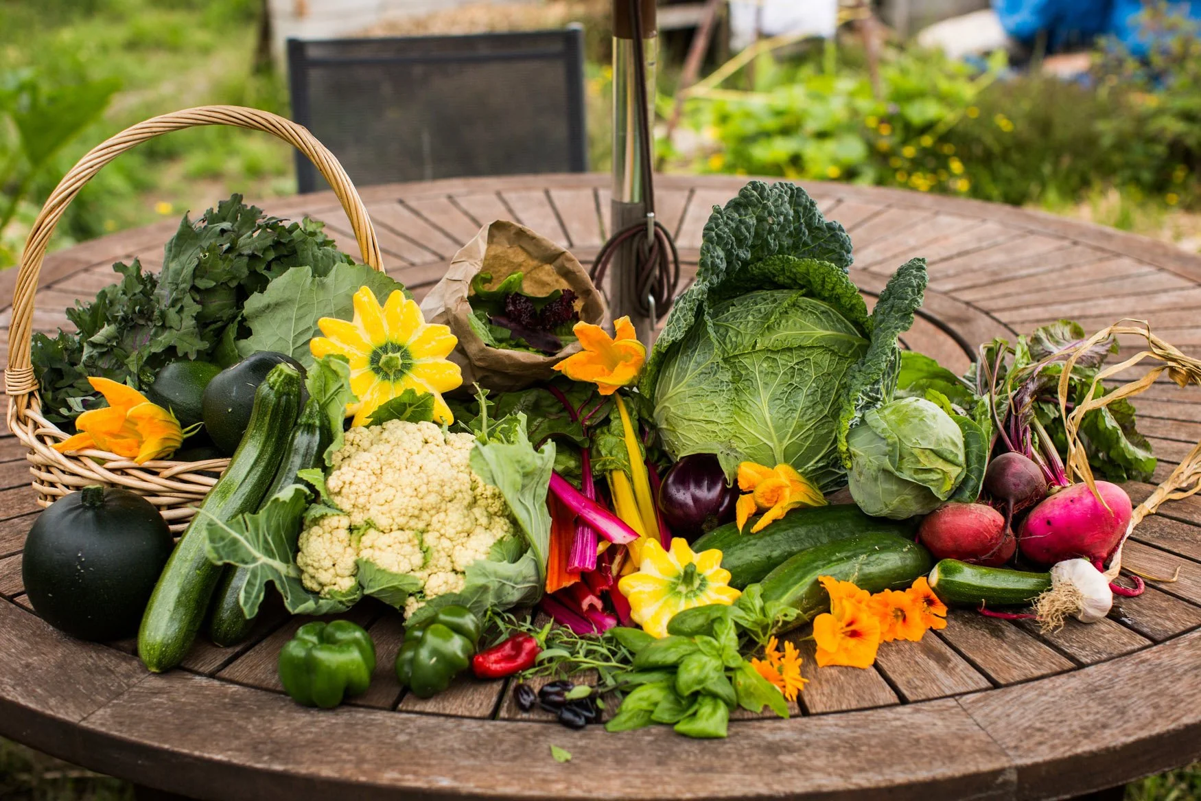 Fresh vegetables and flowers arranged on a round wooden table outdoors, including cauliflower, zucchini, bell peppers, radishes, eggplants, and decorative yellow flowers.