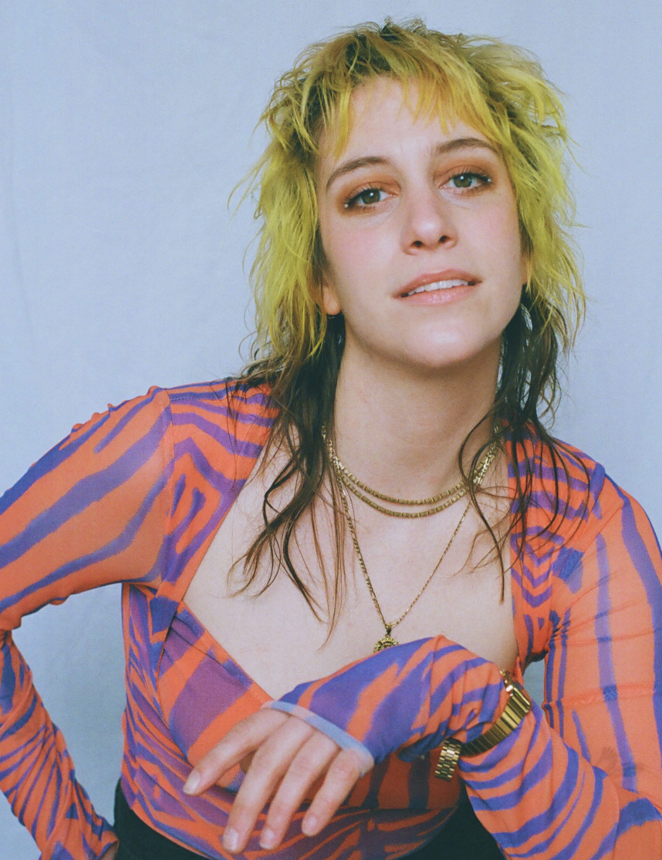 Portrait of a woman with curly, multicolored hair and blue eyes, wearing an orange and purple striped top and layered gold necklaces, against a plain background. Ellen Winter On The Rocks Theatre Edelweiss