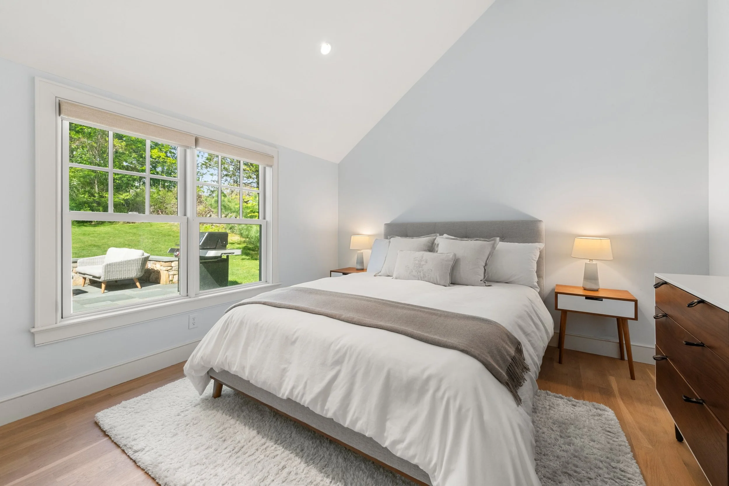 A bedroom with a large bed, nightstands with lamps, a dresser, large window showing a green outdoor scene with trees and grass, and a patio with outdoor furniture and a grill.