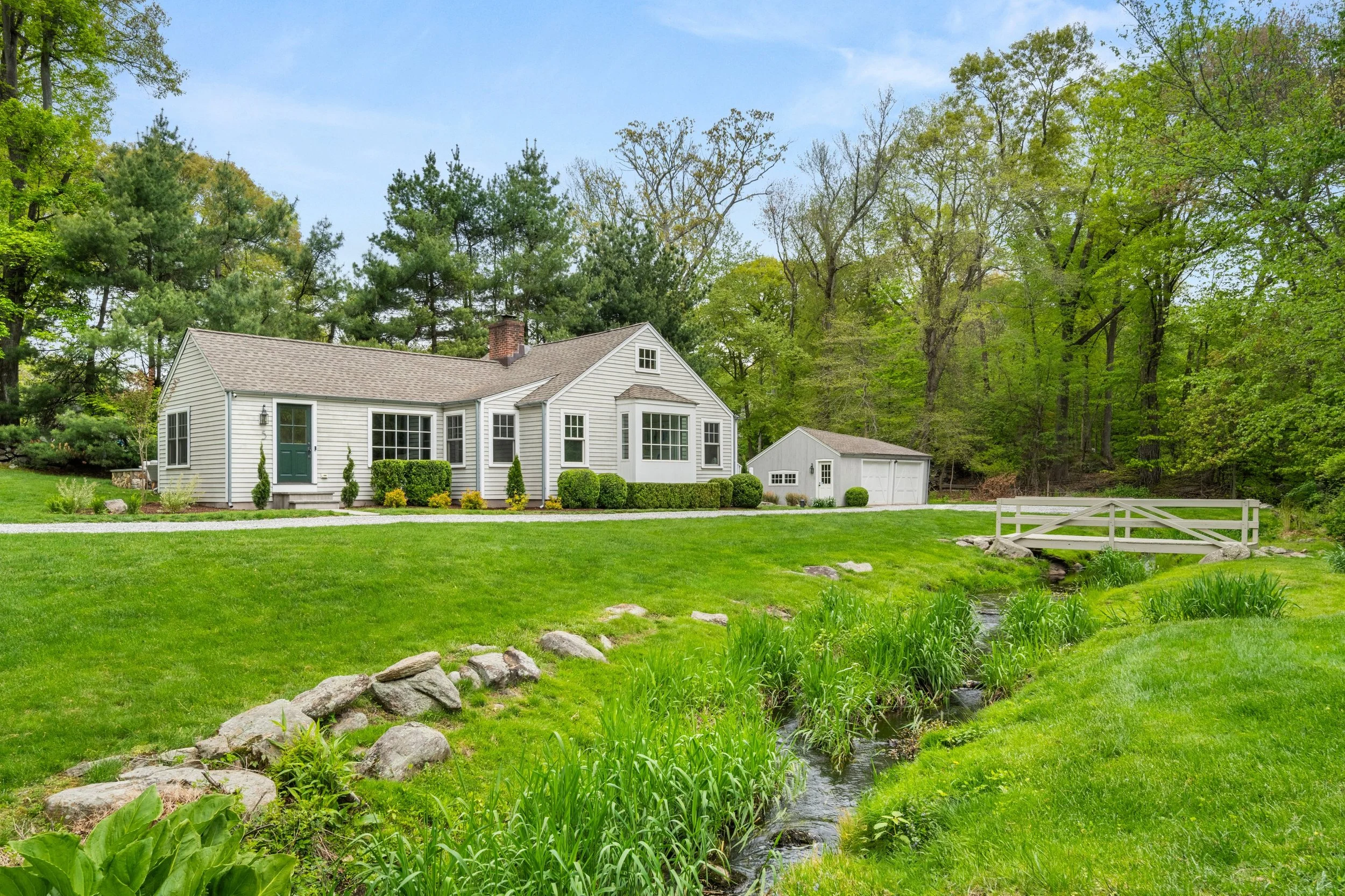 White house with green front door and multiple windows, surrounded by green lawn and garden, with a small stream in the foreground and forest in the background.