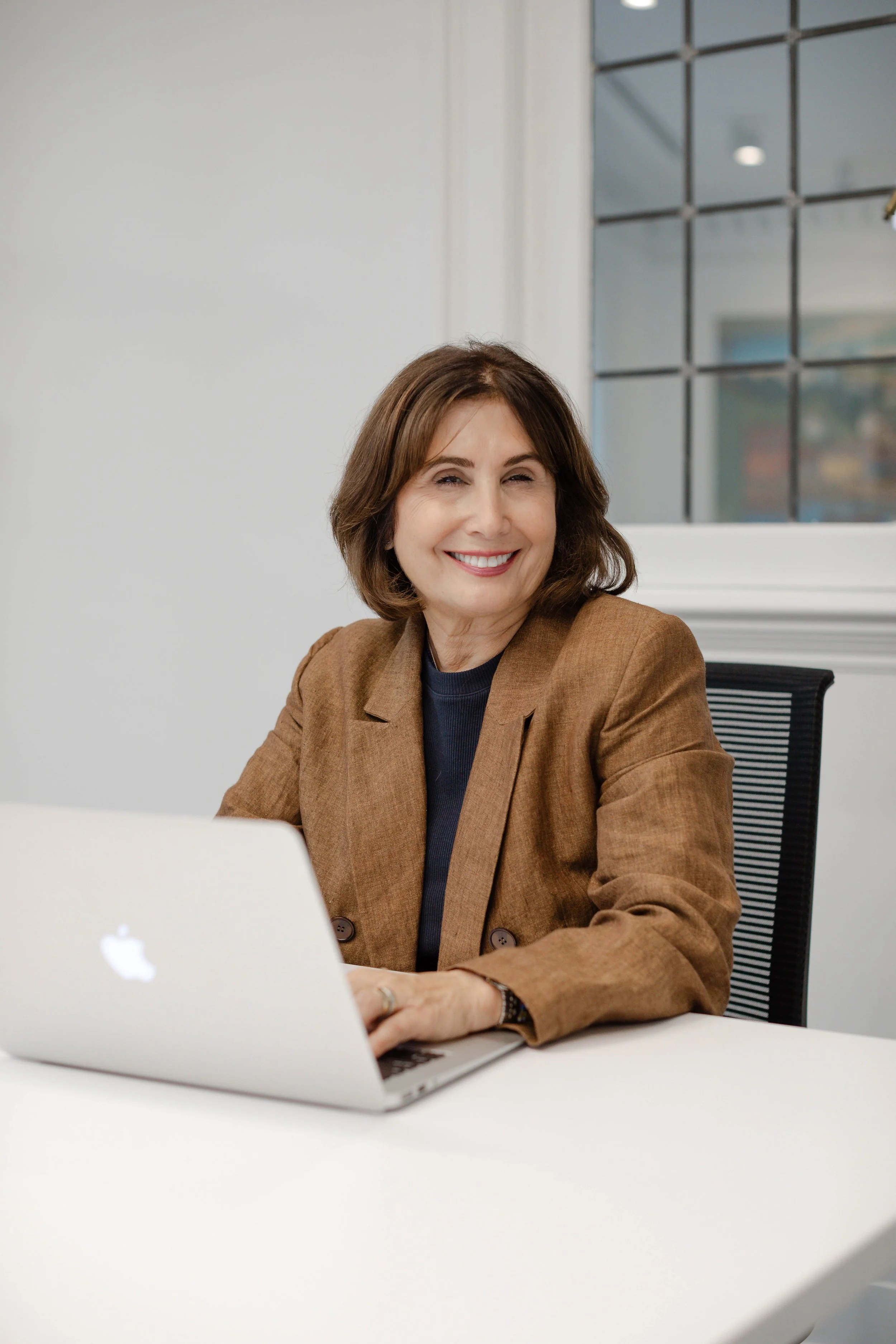 A woman with short brown hair wearing a white suit sitting in a beige chair and smiling, in a minimalistic room with white walls and wooden furniture.
