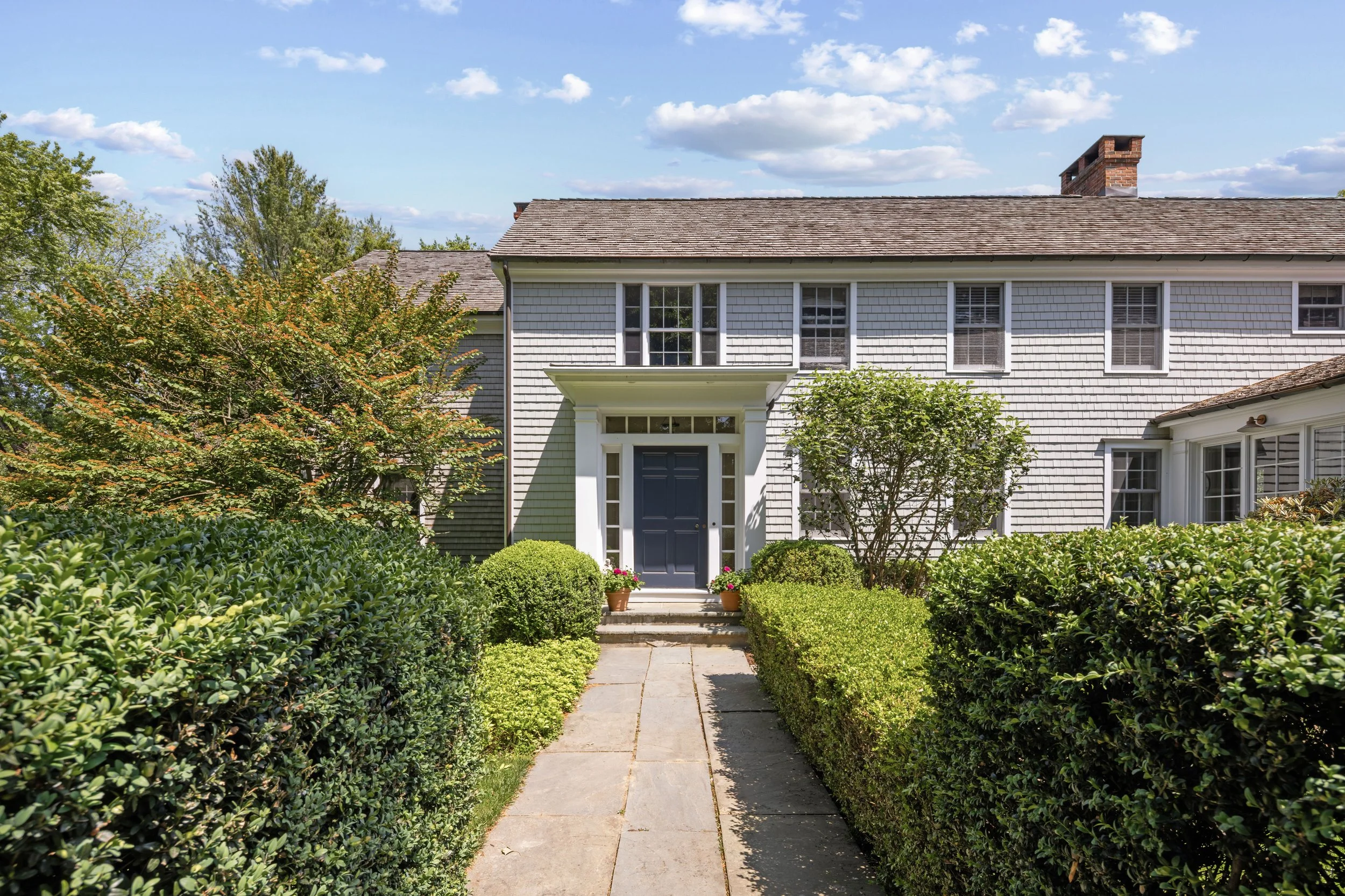 Front view of a two-story house with white siding, a dark blue door, a concrete walkway leading to the front steps, surrounded by shrubs, bushes, and small trees, under a partly cloudy sky.