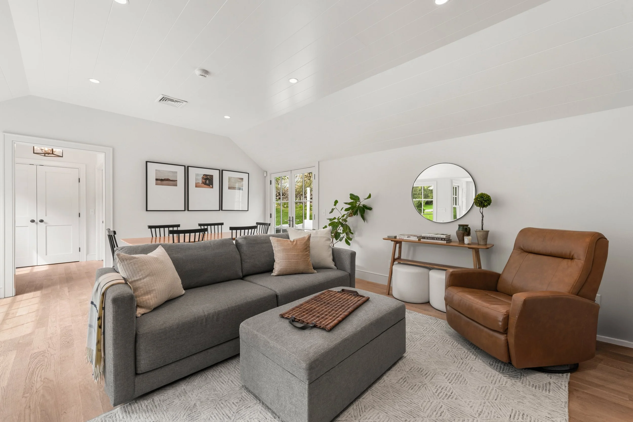 A modern living room with a gray sofa, a brown leather armchair, a side table, a round mirror, and wall art. Large windows and a patterned rug on hardwood floors.