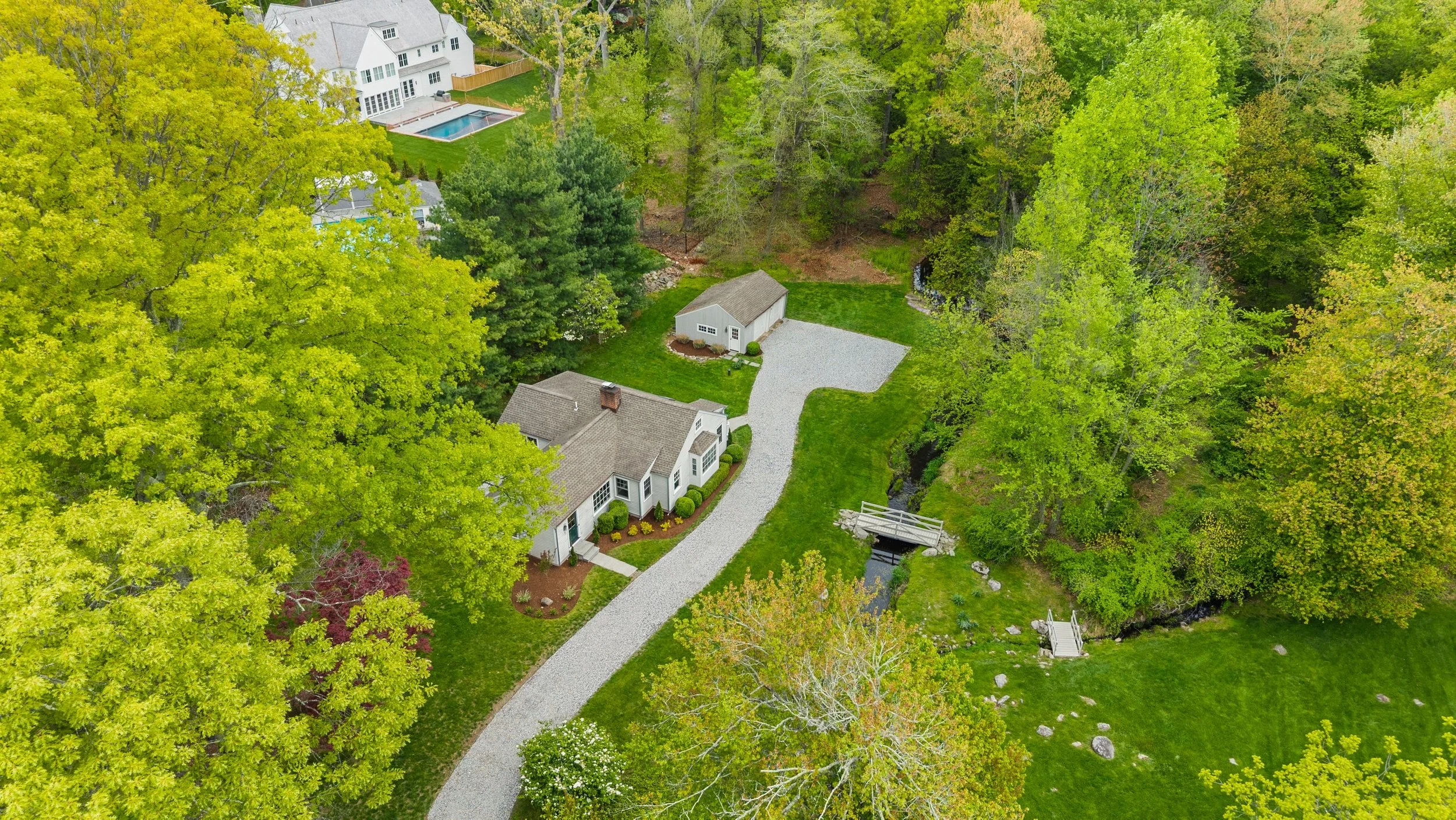Aerial view of a residential house with white siding, gray roof, attached garage, surrounded by lush green trees, a gravel driveway, and a yard with a small stream and bridge.