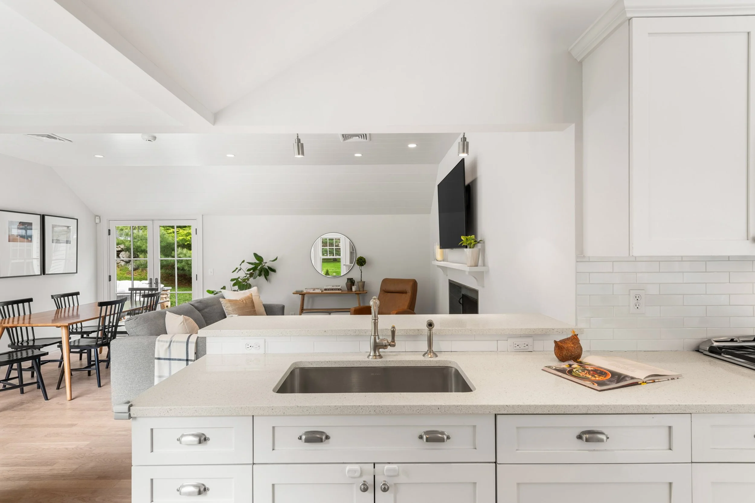 Modern open-concept kitchen with white cabinetry, a large island with a sink, and stainless steel fixtures. In the background, a dining area with a wooden table and black chairs, large windows letting in natural light, and a living room with grey furniture and a fireplace.