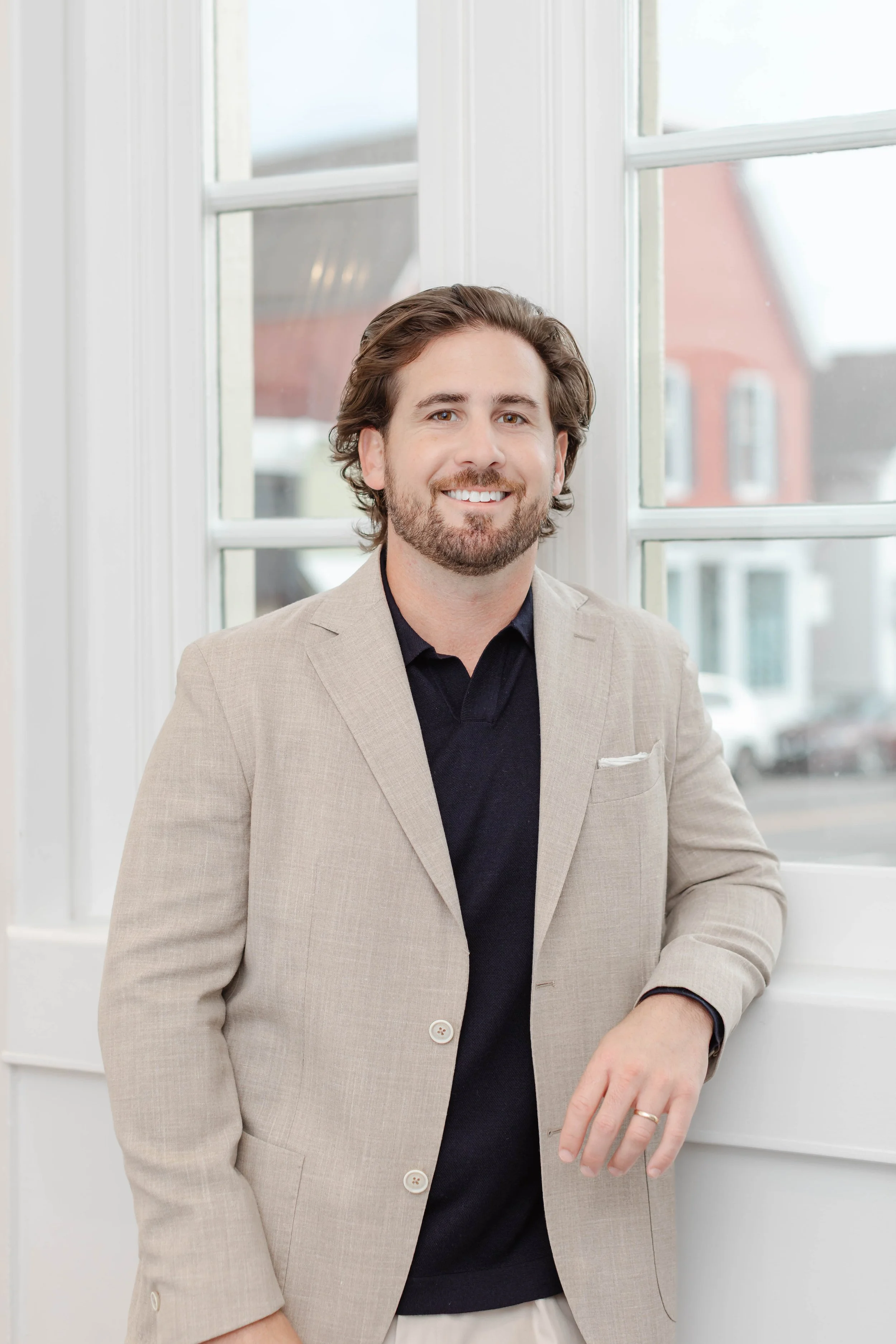 A smiling man with brown hair and a beard, wearing a light-colored checkered blazer, white shirt, and bracelets, standing with arms crossed in front of a white wall near a window.