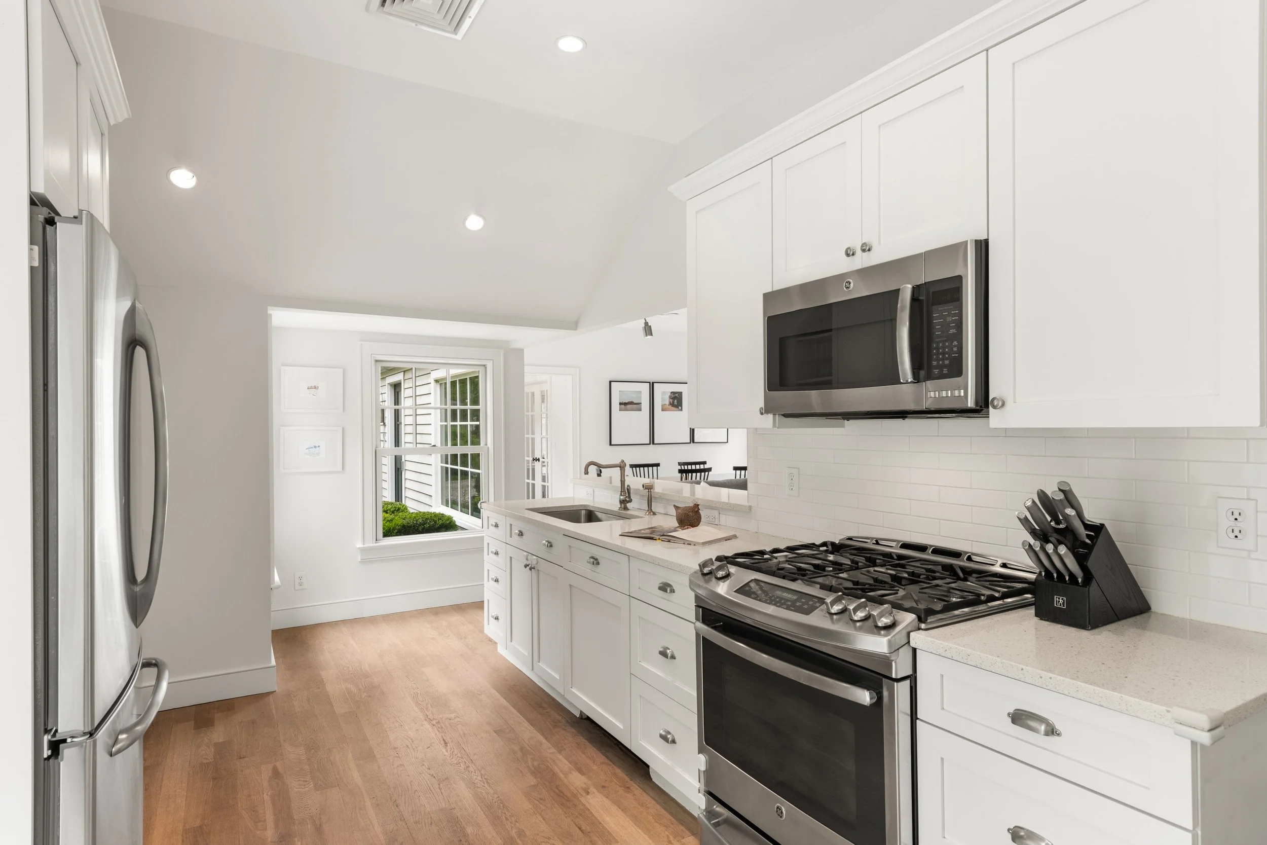 Modern white kitchen with stainless steel appliances, wooden flooring, and large windows.
