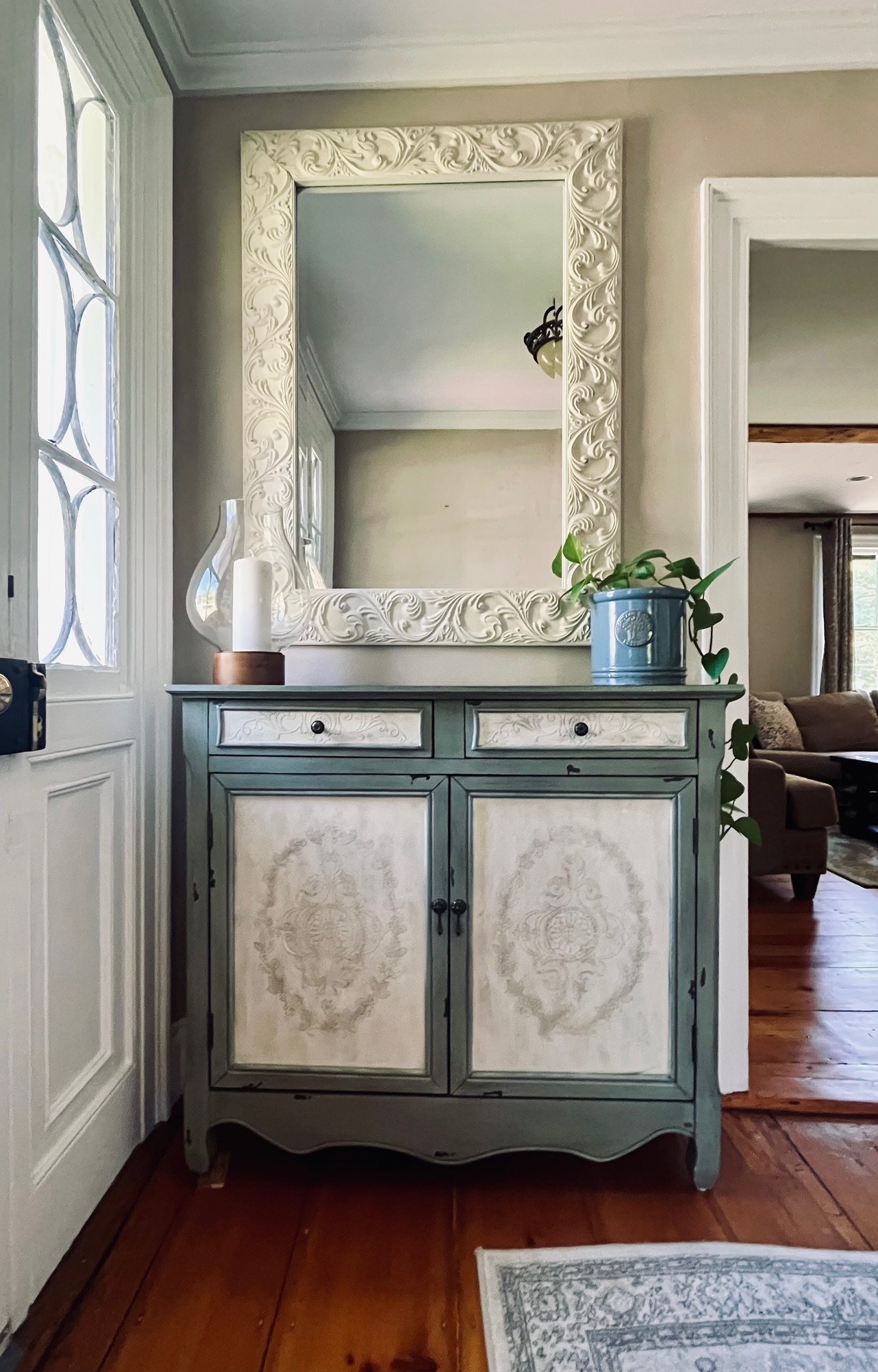 Vintage-style foyer with ornate mirror, decorative cabinet, candleholder, and potted plant.
