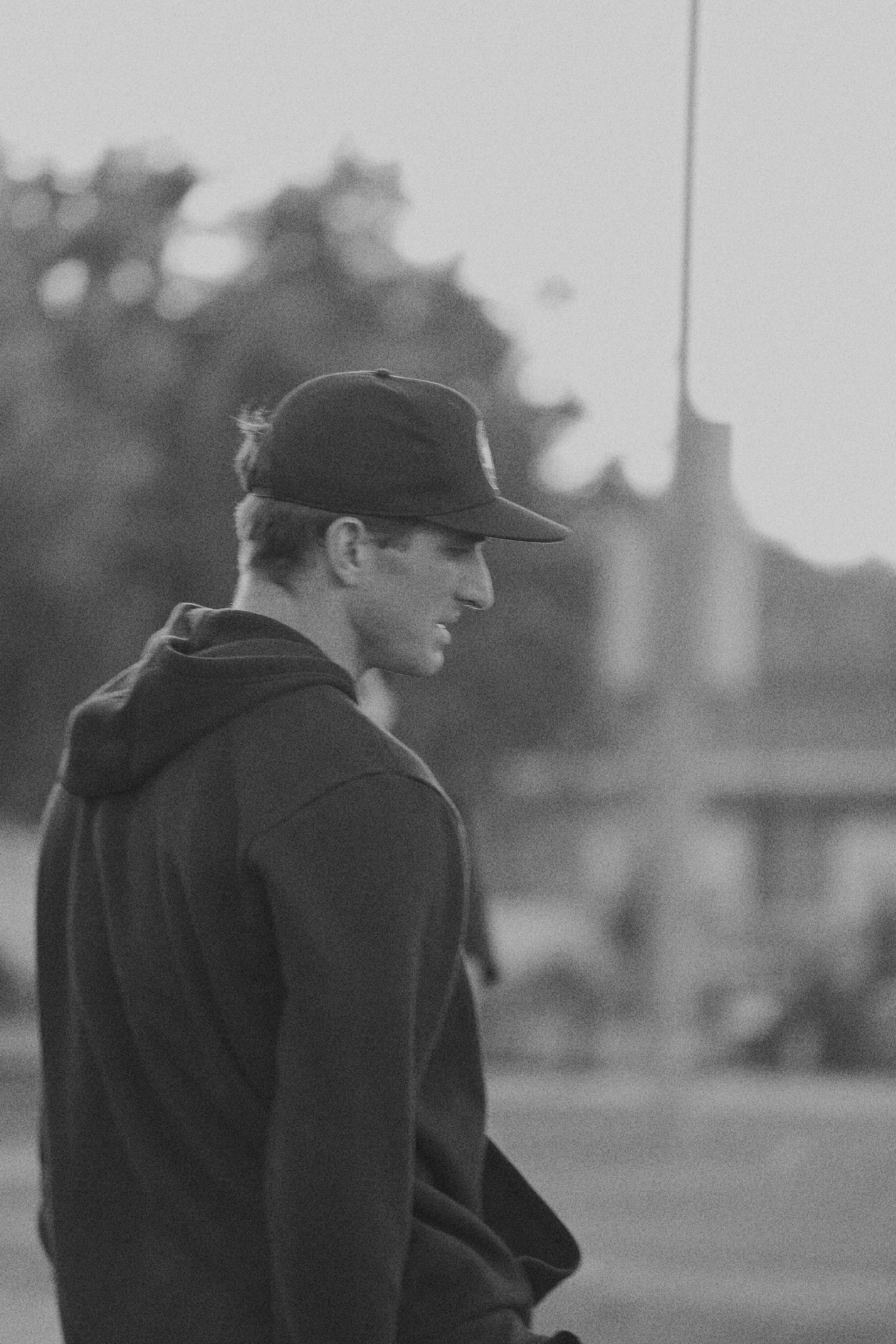 A young man in a baseball cap and hoodie walking outdoors, seen in black and white, with a blurred background of trees and a building.