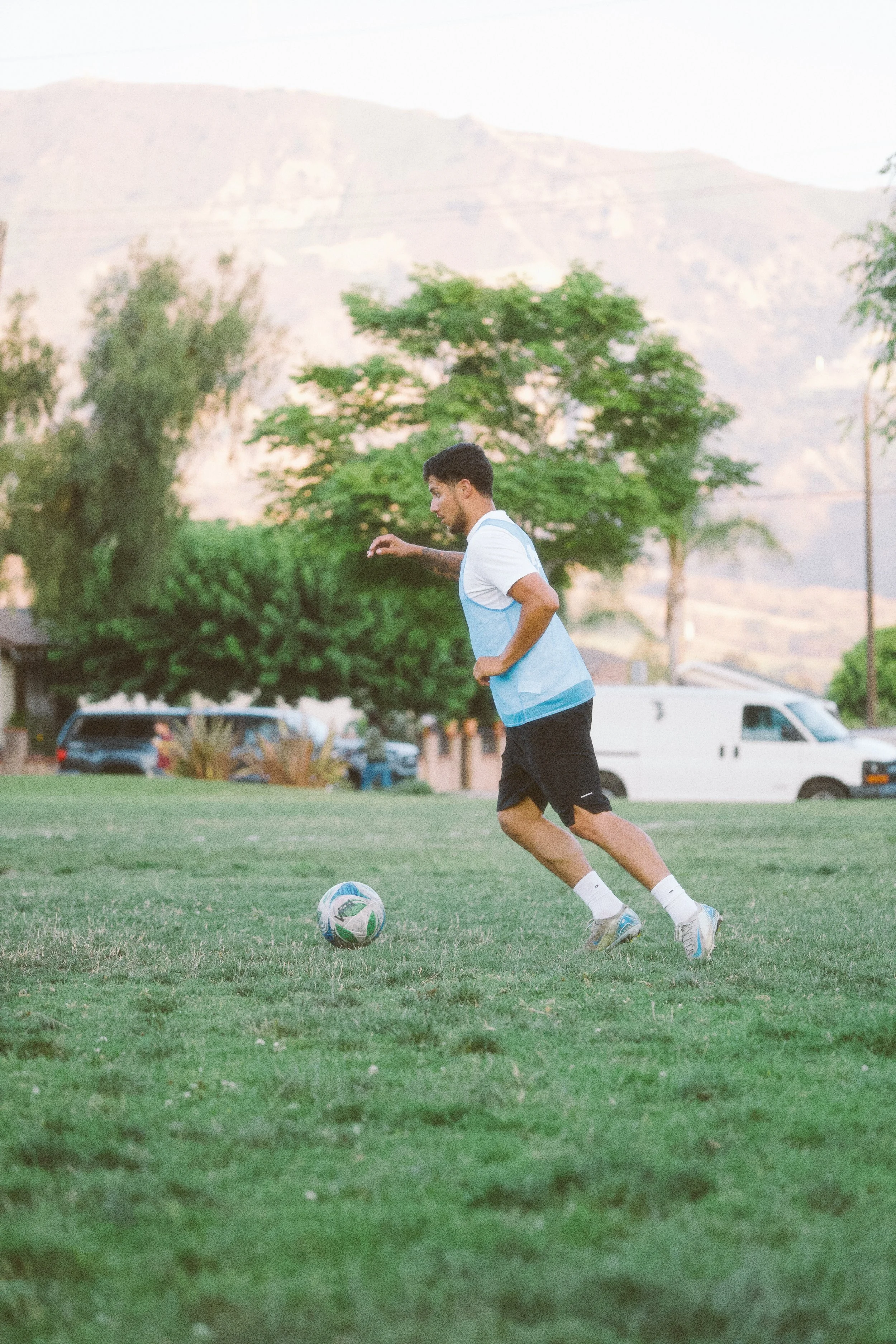 A young man playing soccer outdoors during daytime, running towards a soccer ball on a grassy field with trees and houses in the background.