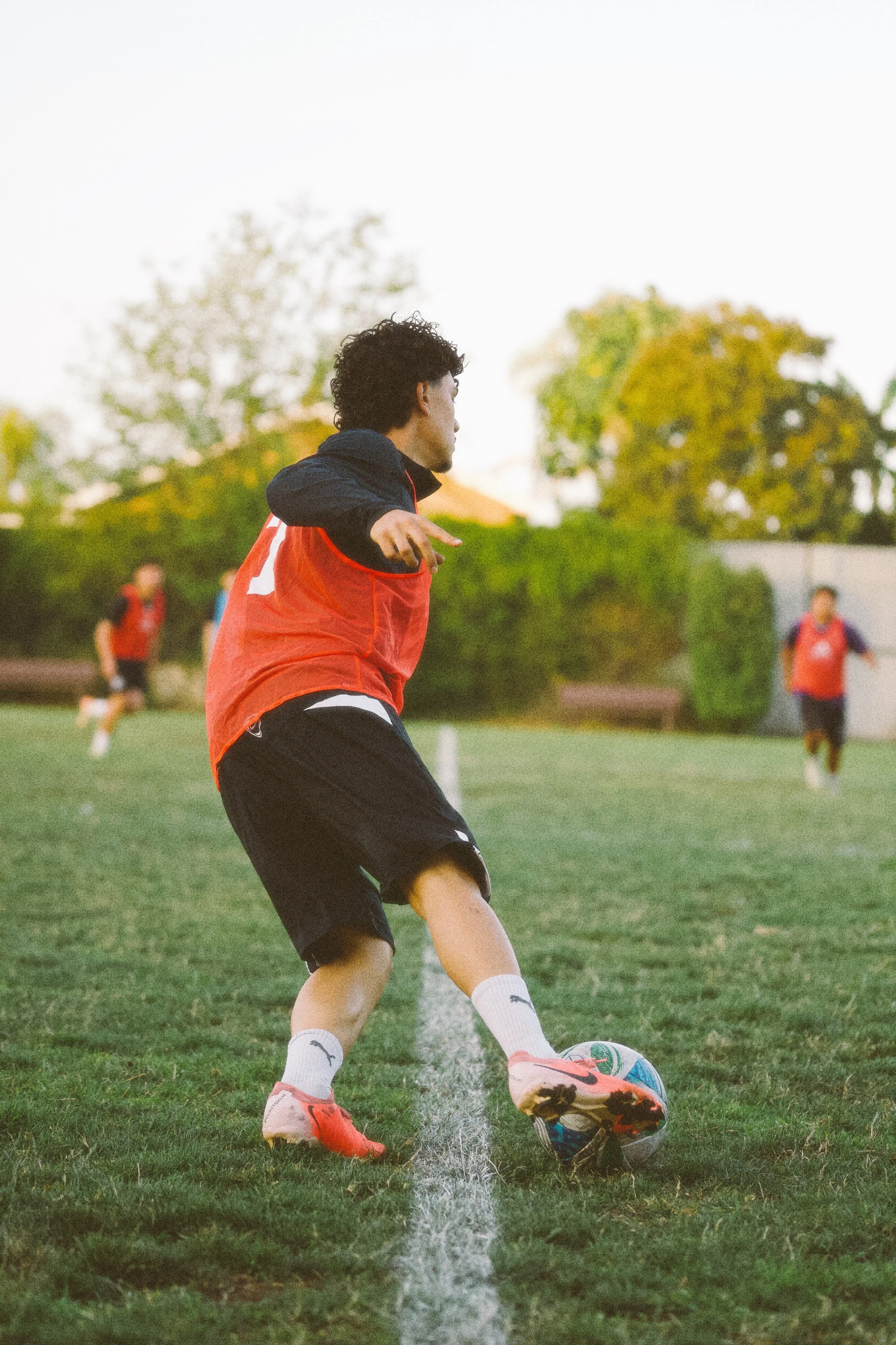 A soccer player in a red vest, black shorts, and red cleats prepares to kick a soccer ball on a grassy field with other players in the background.
