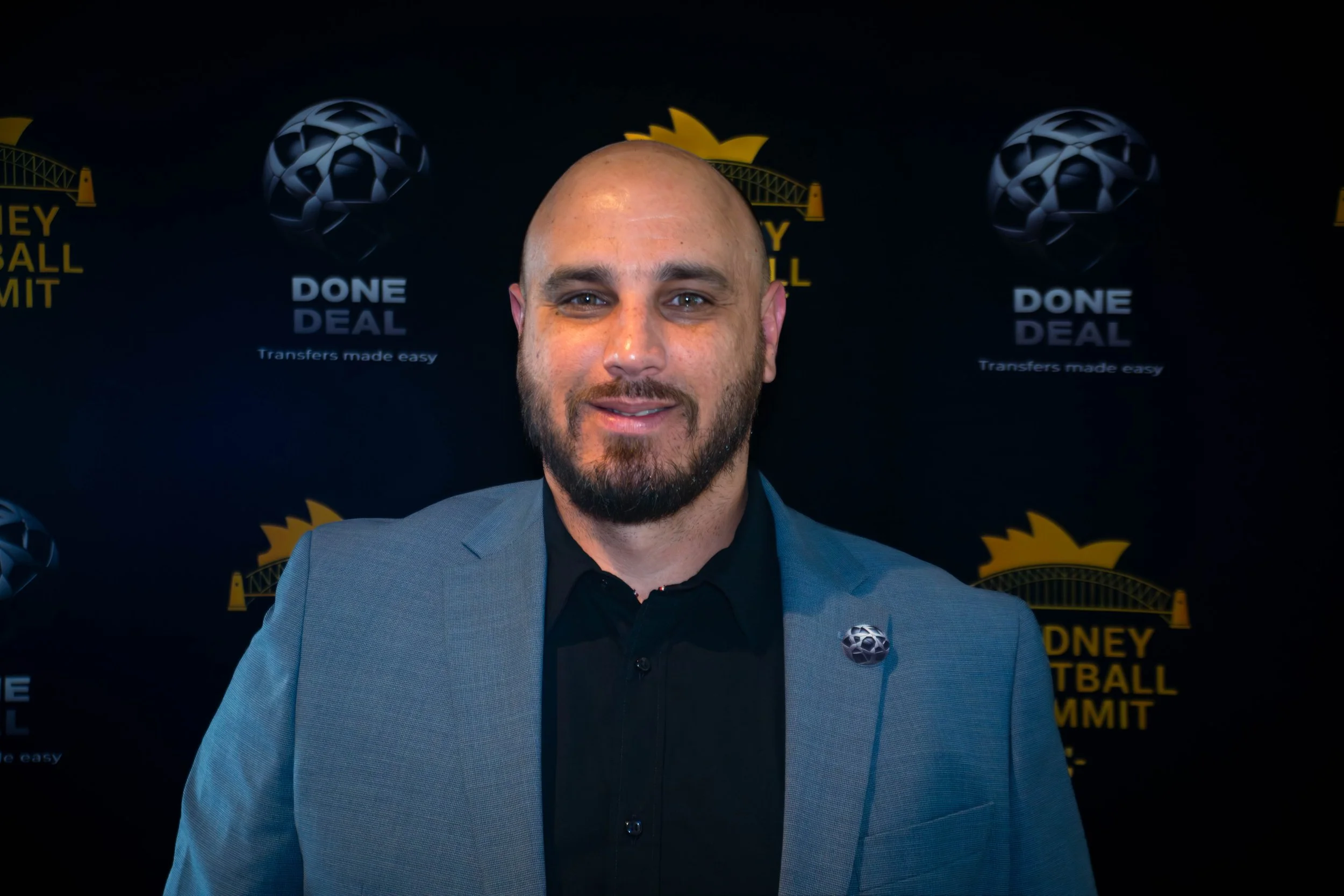 A man with a beard and a shaved head wearing a gray blazer and black shirt, standing in front of a black backdrop with yellow and white logos and the text 'DONE DEAL Transfers made easy' and 'MONEY FOOTBALL SUMMIT'.