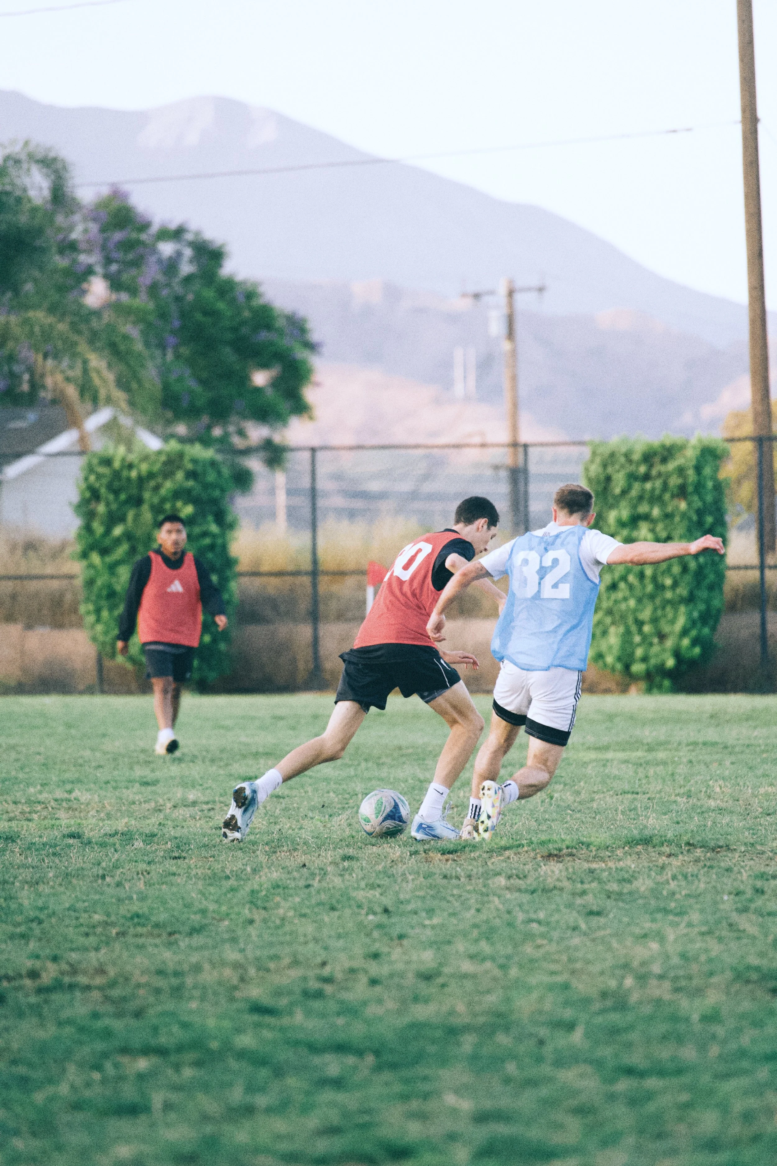 Two boys playing soccer on a grass field with a mountain and trees in the background, one in a red jersey and the other in a light blue jersey.