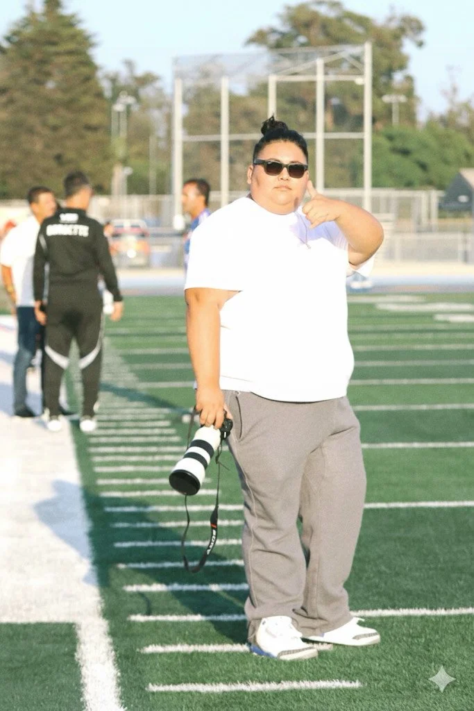Person with sunglasses holding a camera on a football field, with other people in the background.