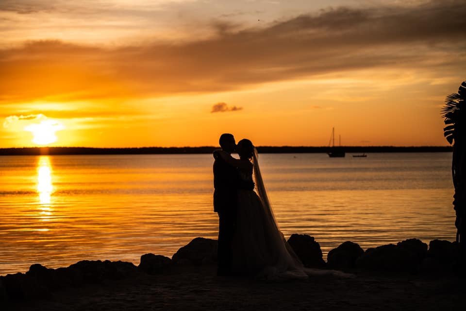 Silhouette of a couple hugging by the water during a vibrant sunset with boats in the distance.