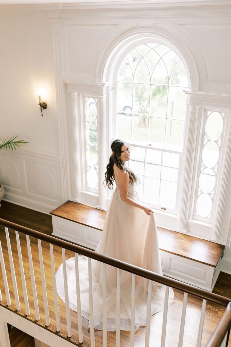 Bride in a white wedding dress standing on a staircase in front of a large arched window, smiling and looking over her shoulder.