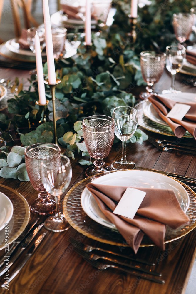 Elegant dining table set for a formal event, featuring gold-rimmed plates, blush pink napkins, crystal glasses, and a lush greenery centerpiece with pink candles.
