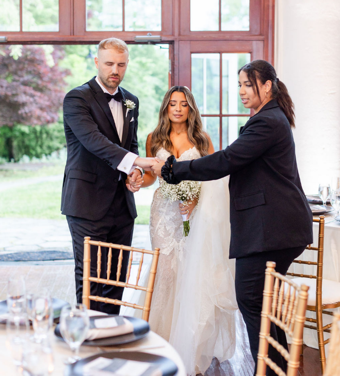 A wedding ceremony with a bride and groom holding hands while an officiant conducts the ceremony inside a room with large windows and scenic outdoor views.