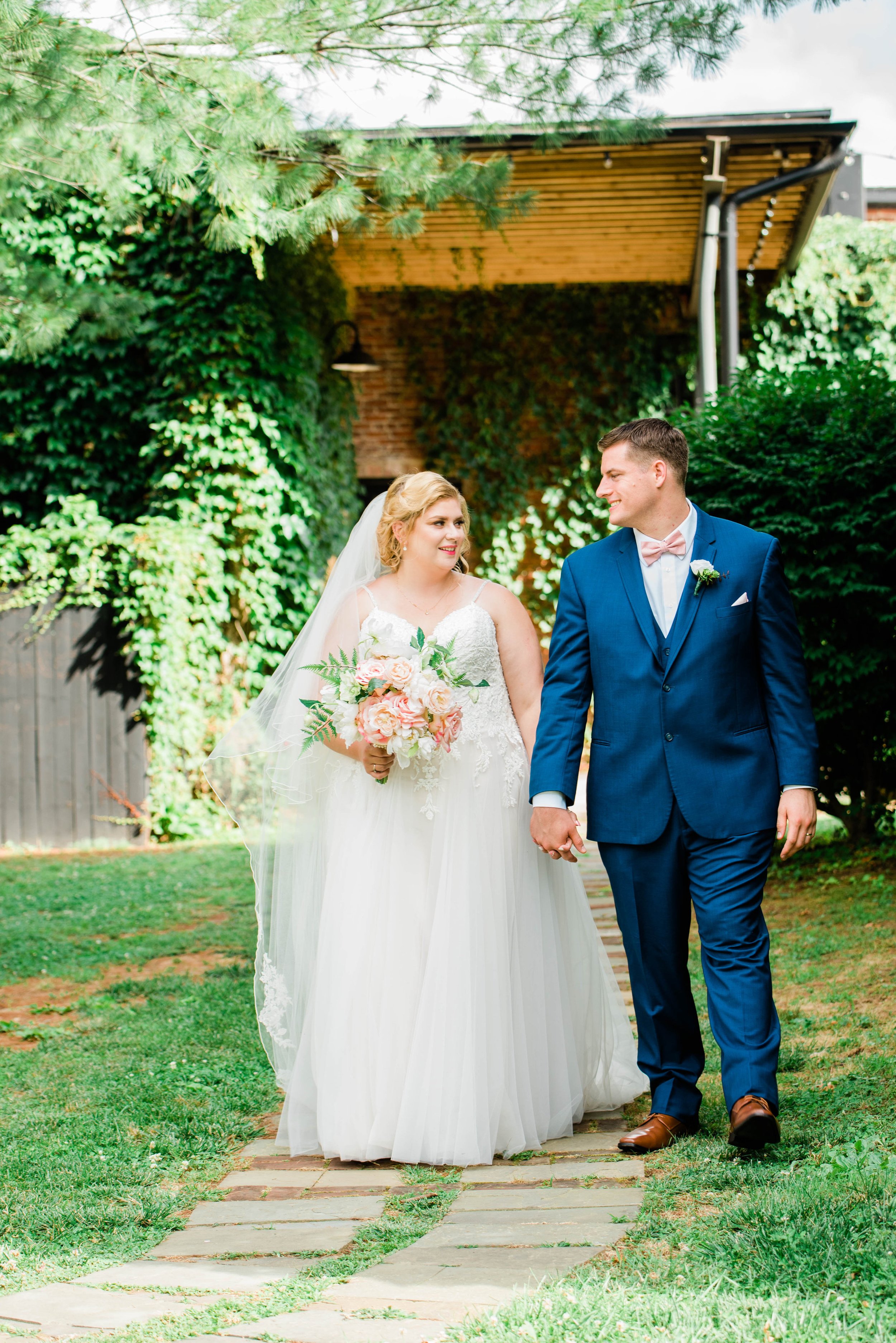 A bride and groom walking hand in hand outdoors on a garden path during their wedding, surrounded by green trees and foliage.