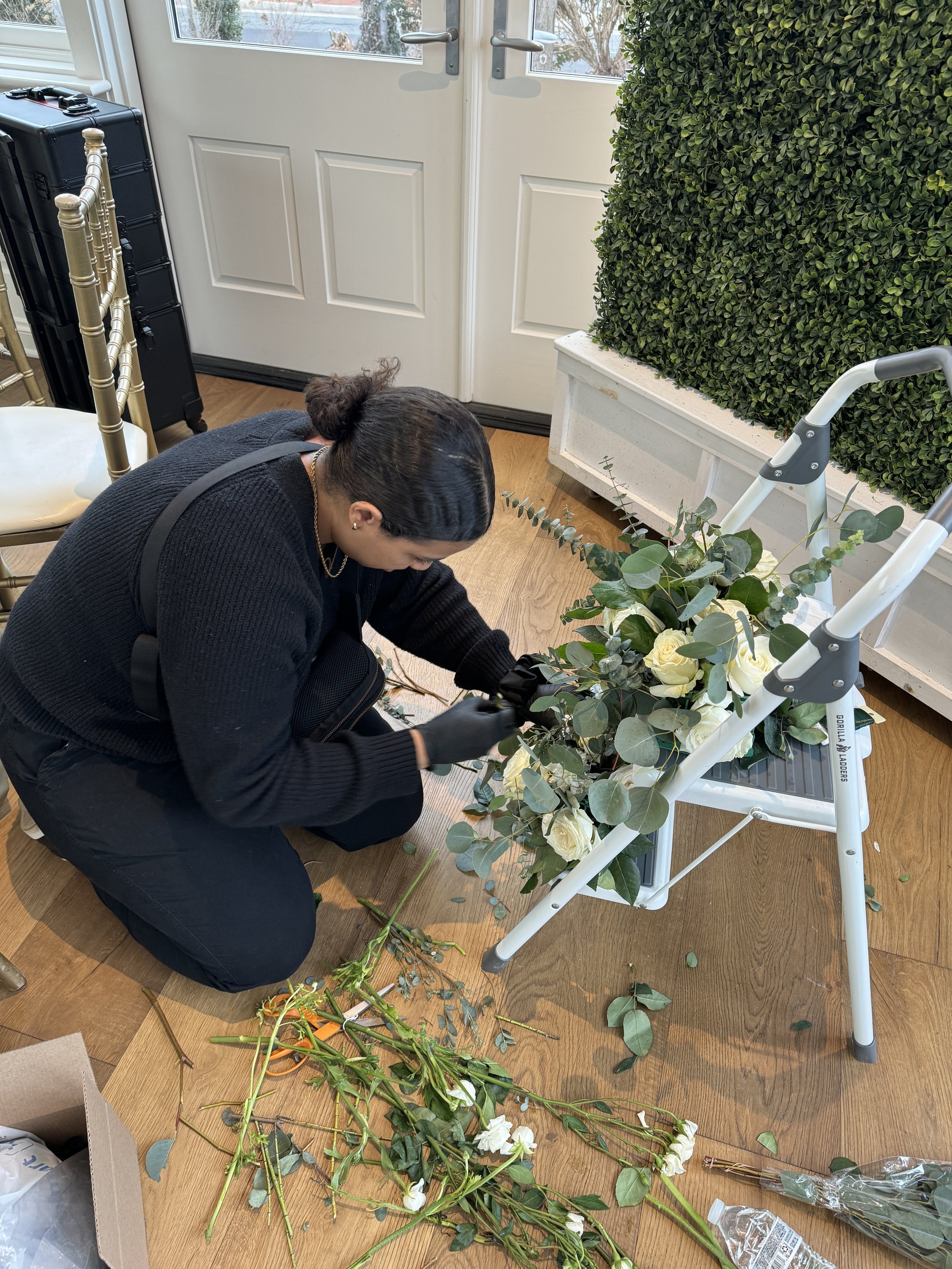 A woman arranging white roses and greenery on a white step ladder indoors near a door with glass window panels.