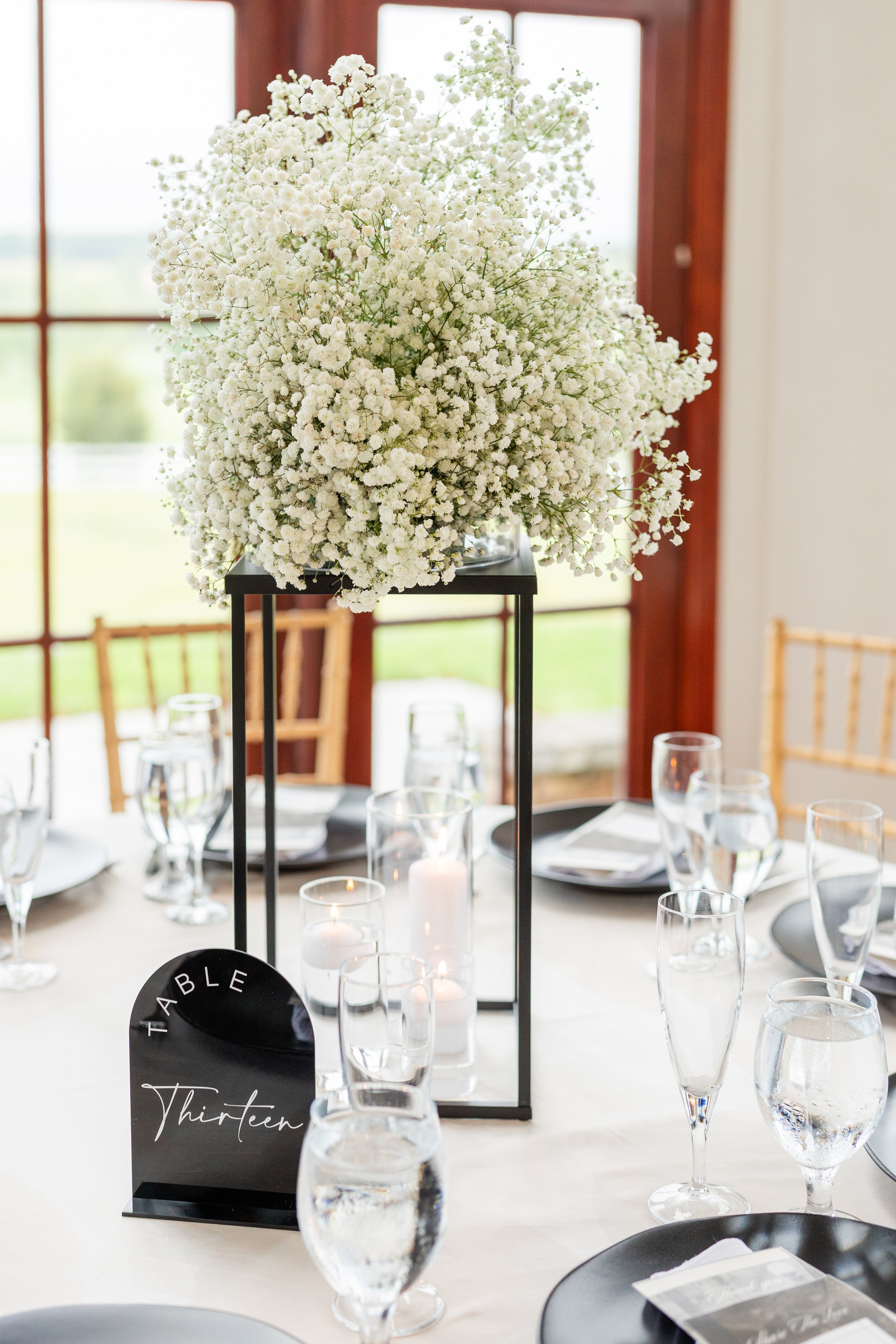 Elegant table centerpiece with white baby's breath flowers on a black metal stand, surrounded by water glasses, candles, and place settings, at a bright indoor event space.