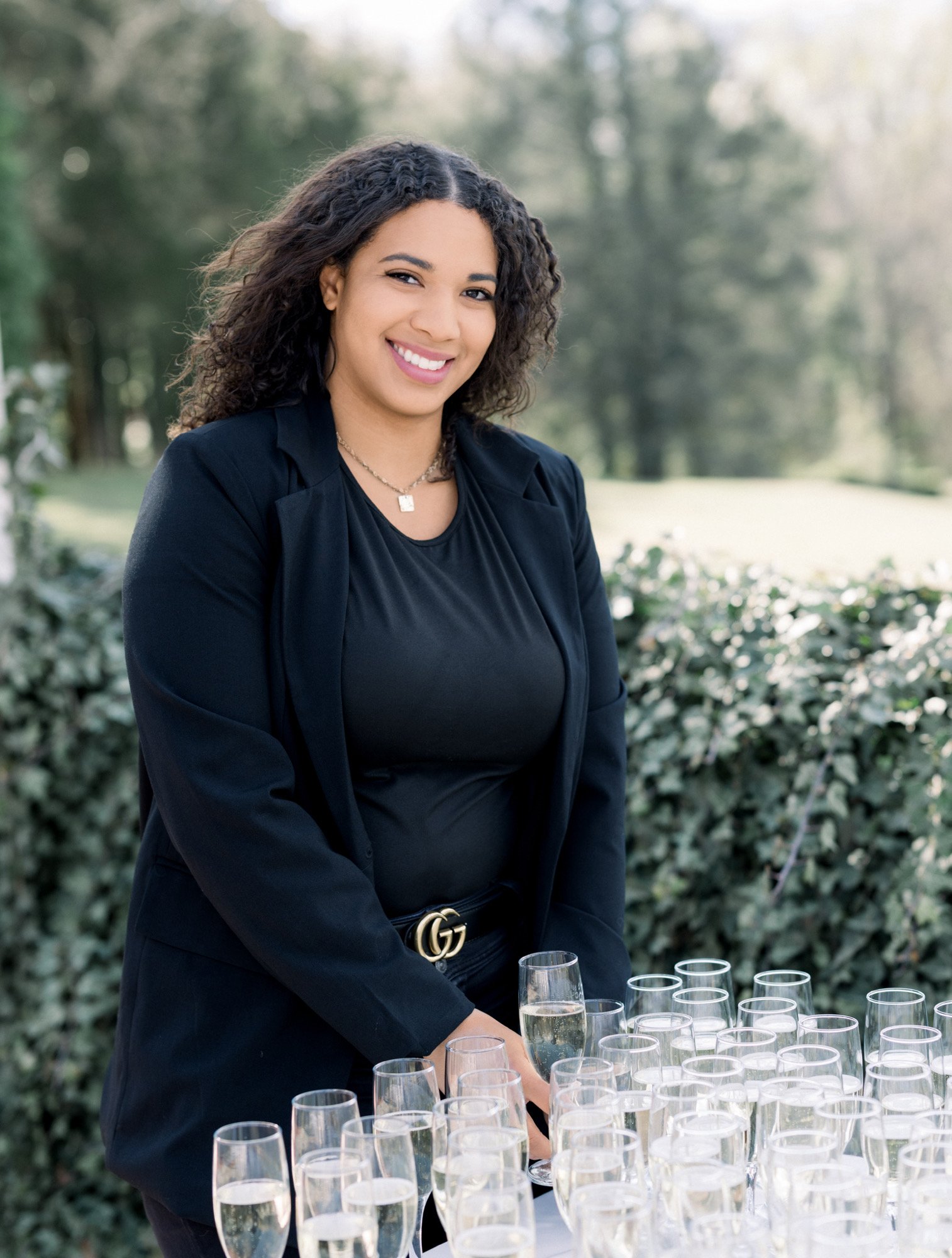 A woman in black attire standing outdoors beside a table with numerous champagne glasses filled with champagne, smiling at the camera.