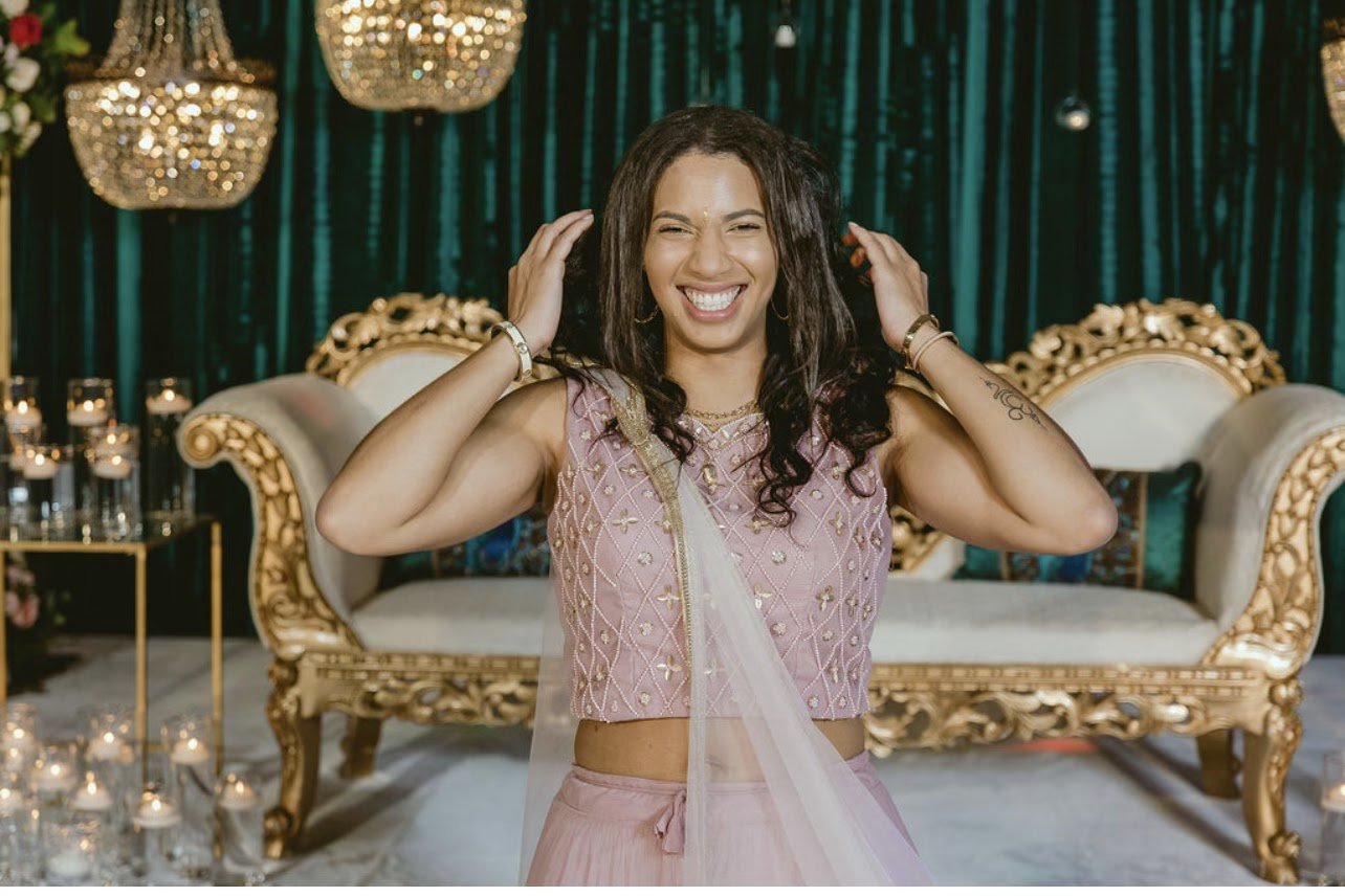A woman in traditional South Asian attire smiling at a celebratory event with ornate gold furniture and decorative lighting.