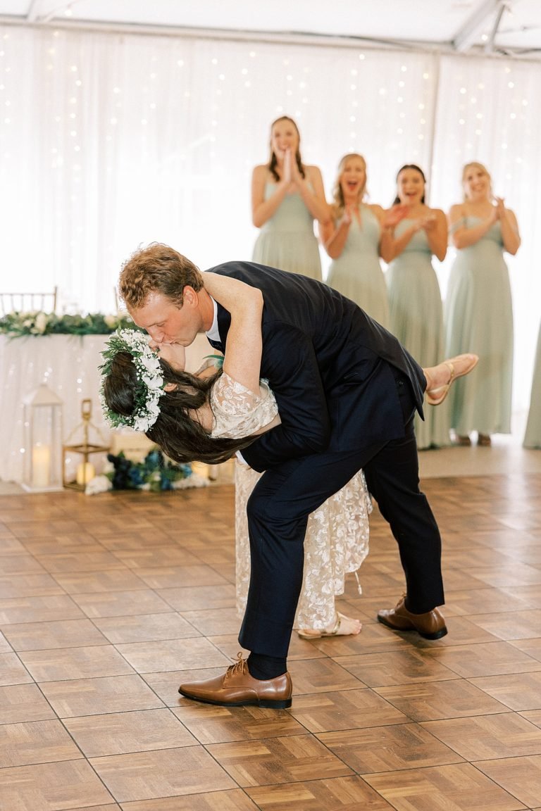 A bride and groom share a kiss on the dance floor during their wedding reception, with bridesmaids clapping and smiling in the background.