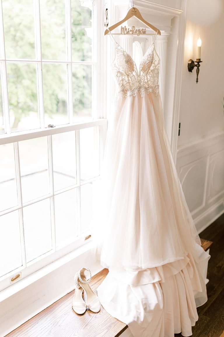 White wedding dress hanging from a hanger on a window frame with labeled name 'Unbekannt'; white wedding shoes with ankle straps placed on the wooden floor.