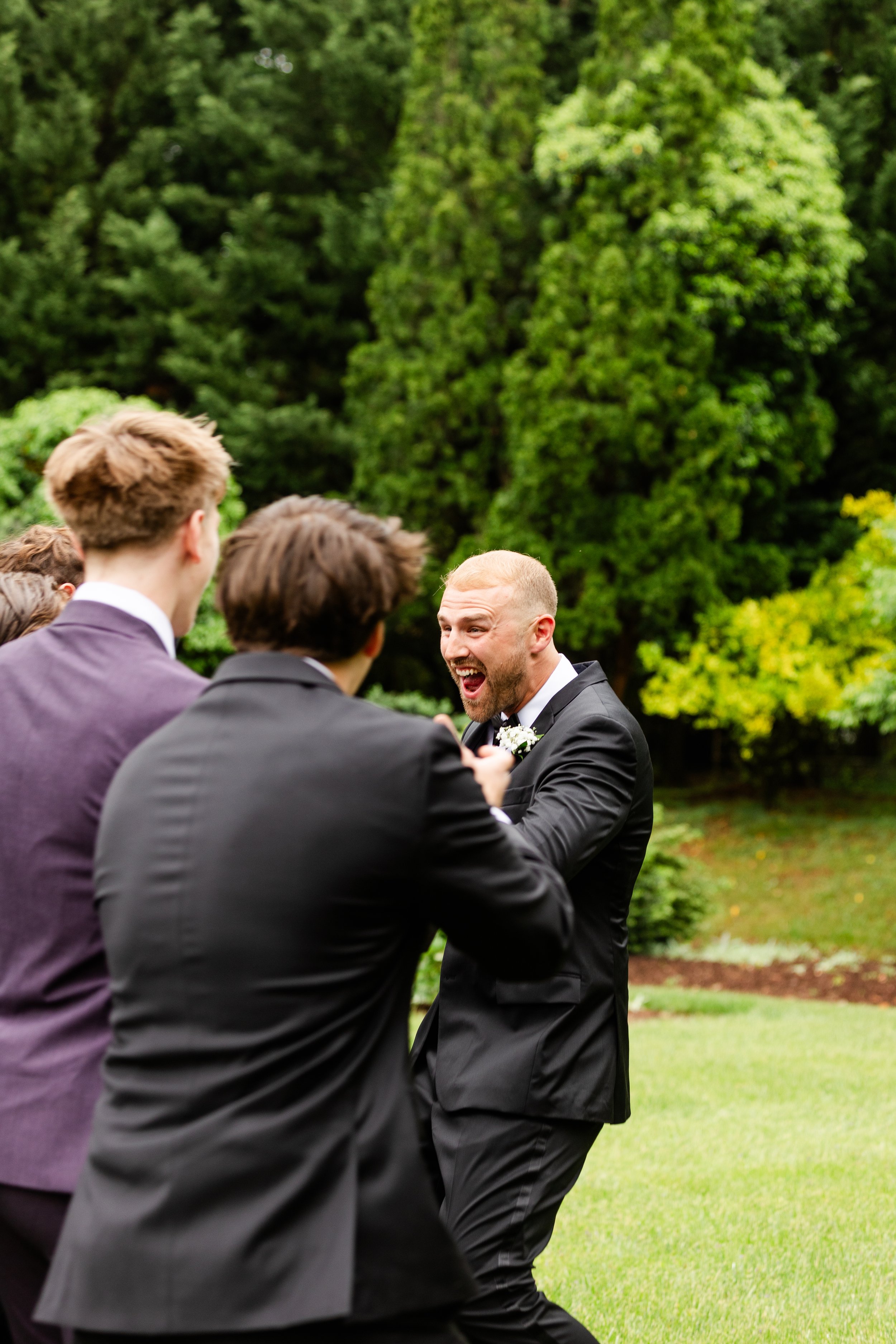 A group of men in suits enjoying a lively moment outdoors in a lush green park during a wedding celebration.