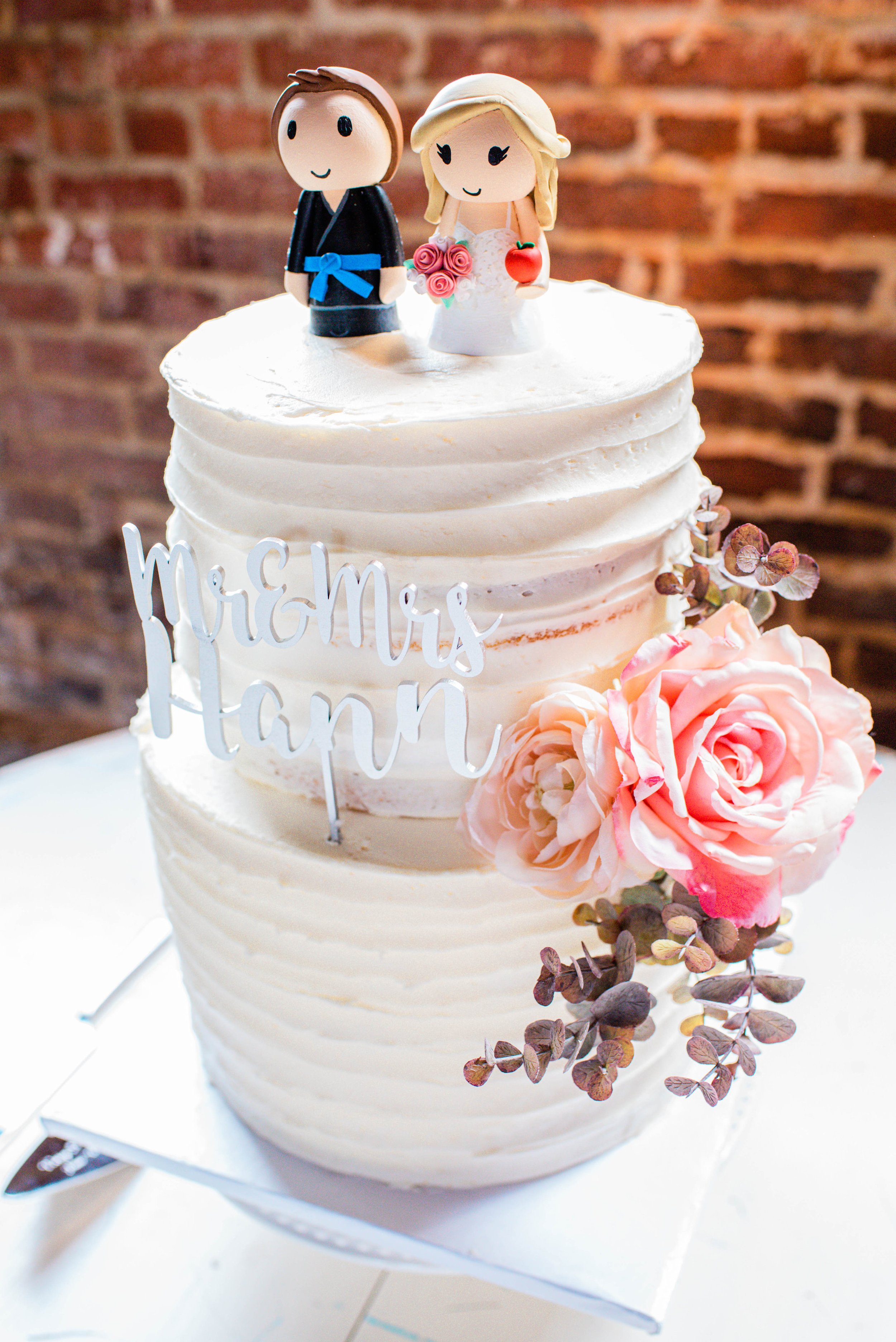 A three-tier wedding cake decorated with pink roses, greenery, a "Mr & Mrs tan" cake topper, and two fondant figurines of a bride and groom on top. The groom wears a tuxedo with a blue sash, and the bride wears a wedding dress and holds pink roses an
