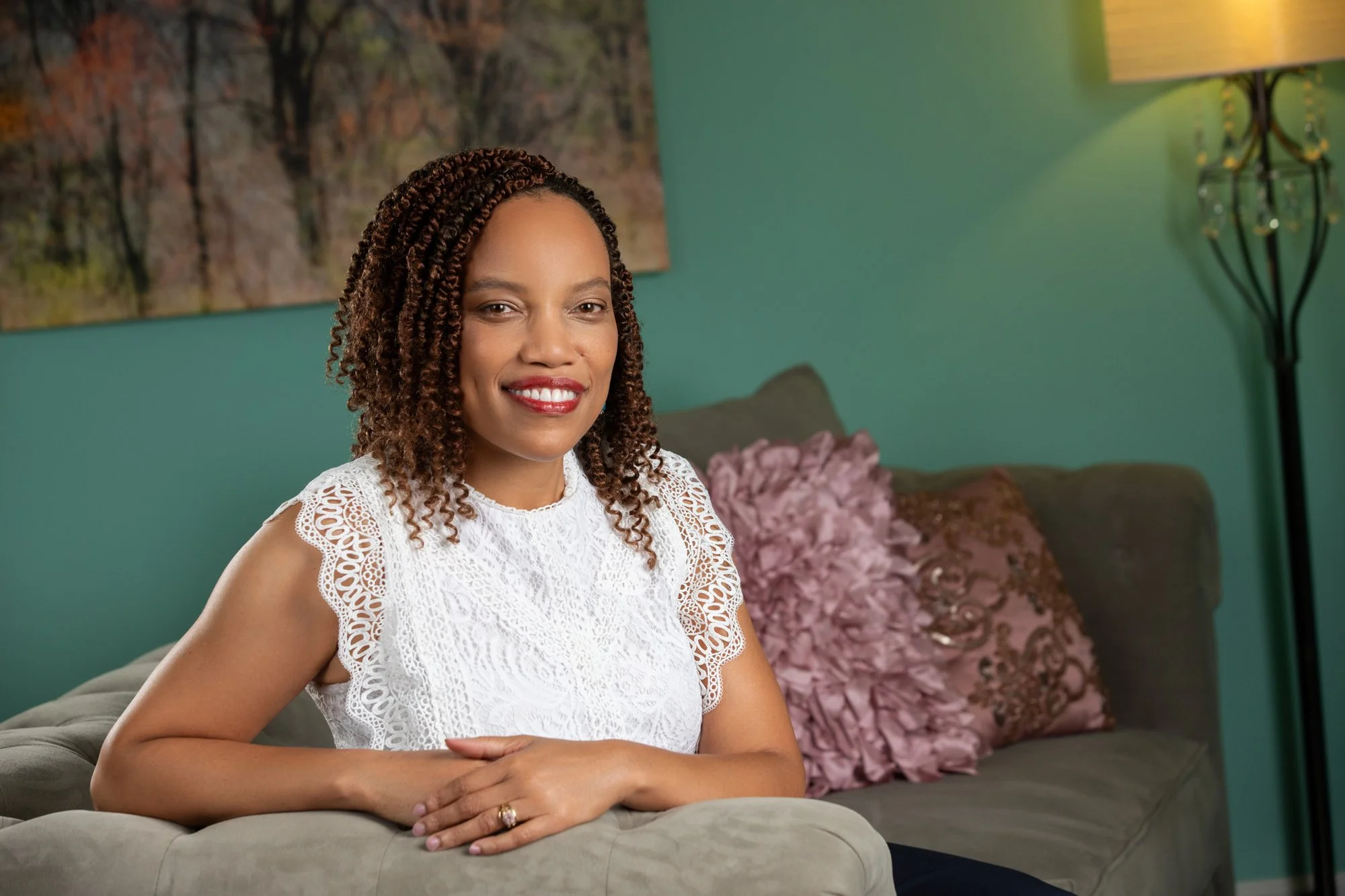 Author Antoinette H. Jones sitting on a couch with decorative pillows, wearing a white lace top, in a living room with teal walls and a lamp.