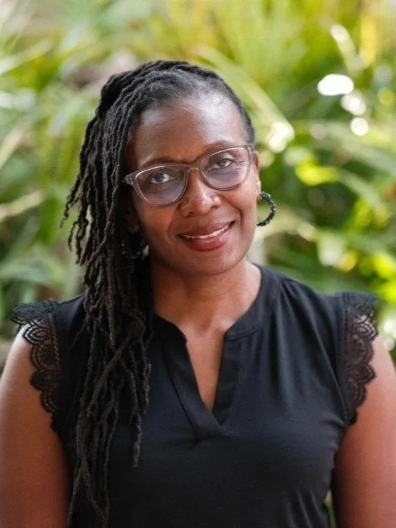 A woman with glasses and long dreadlocks smiling outdoors with green foliage in the background.