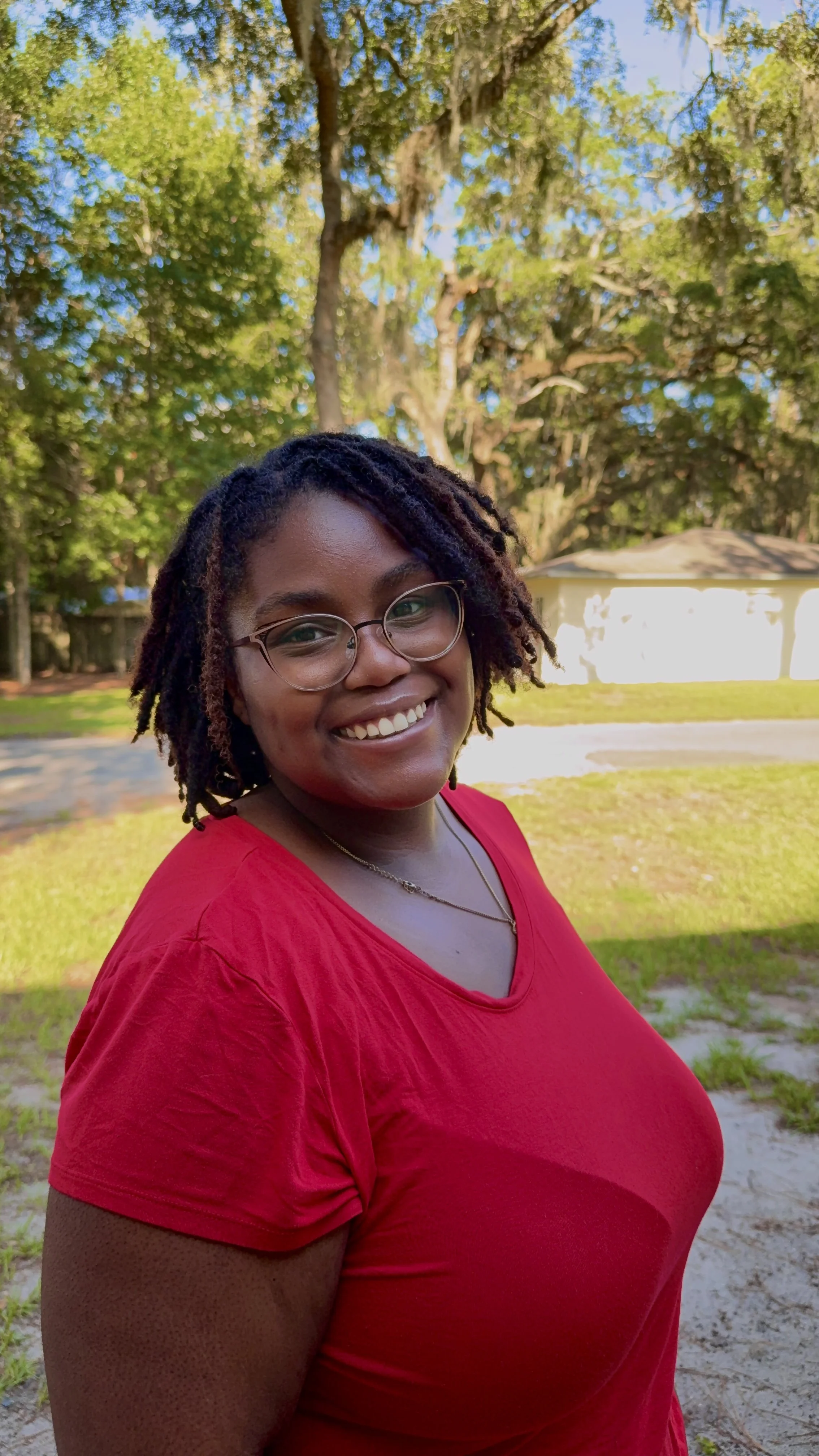 Lacorya, with black and brown locs and rose gold glasses, is wearing a red shirt and smiling in front of a blurry background of a house and trees.