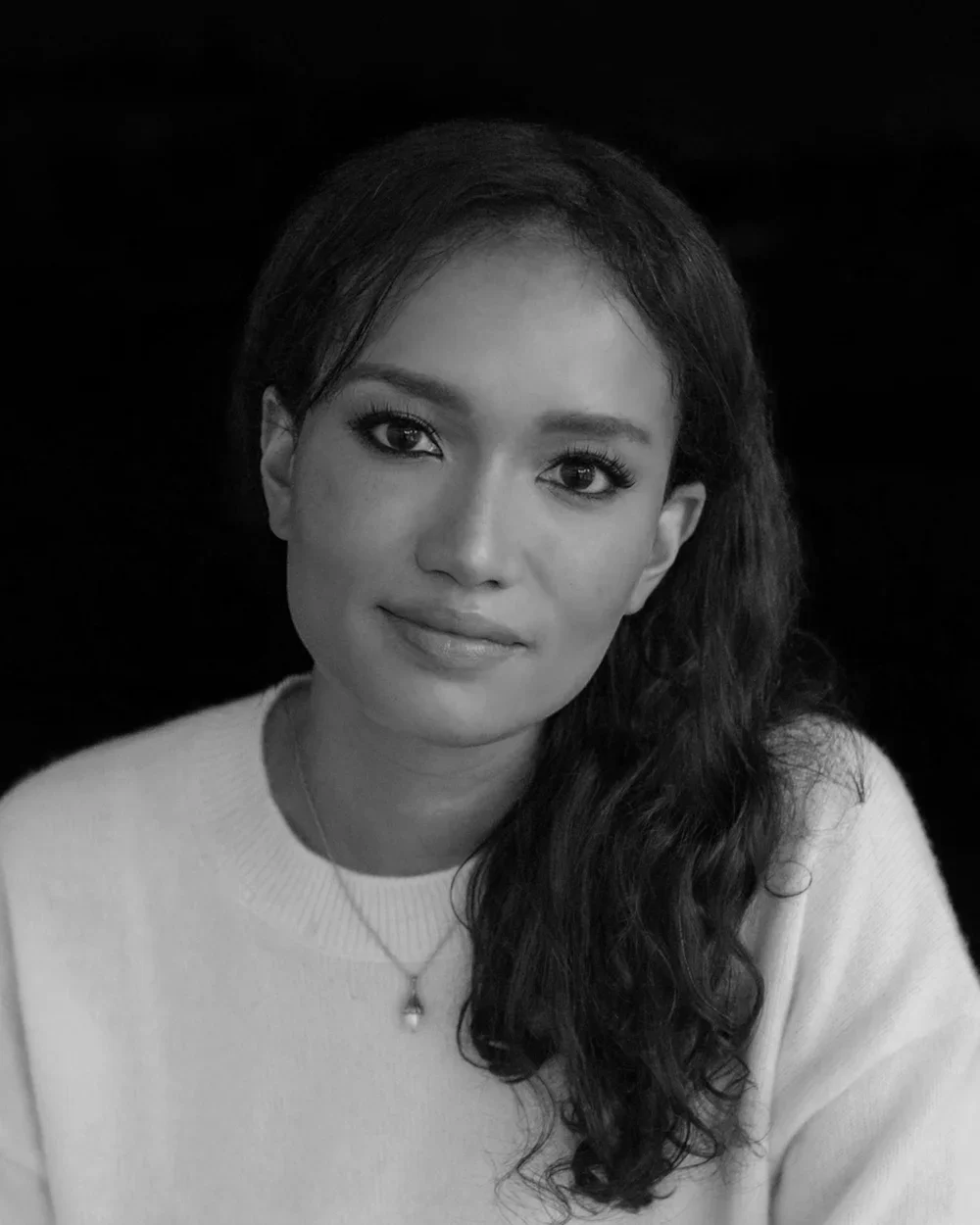 Portrait of a woman with long, wavy hair and makeup, wearing a light-colored top and a necklace, against a black background.