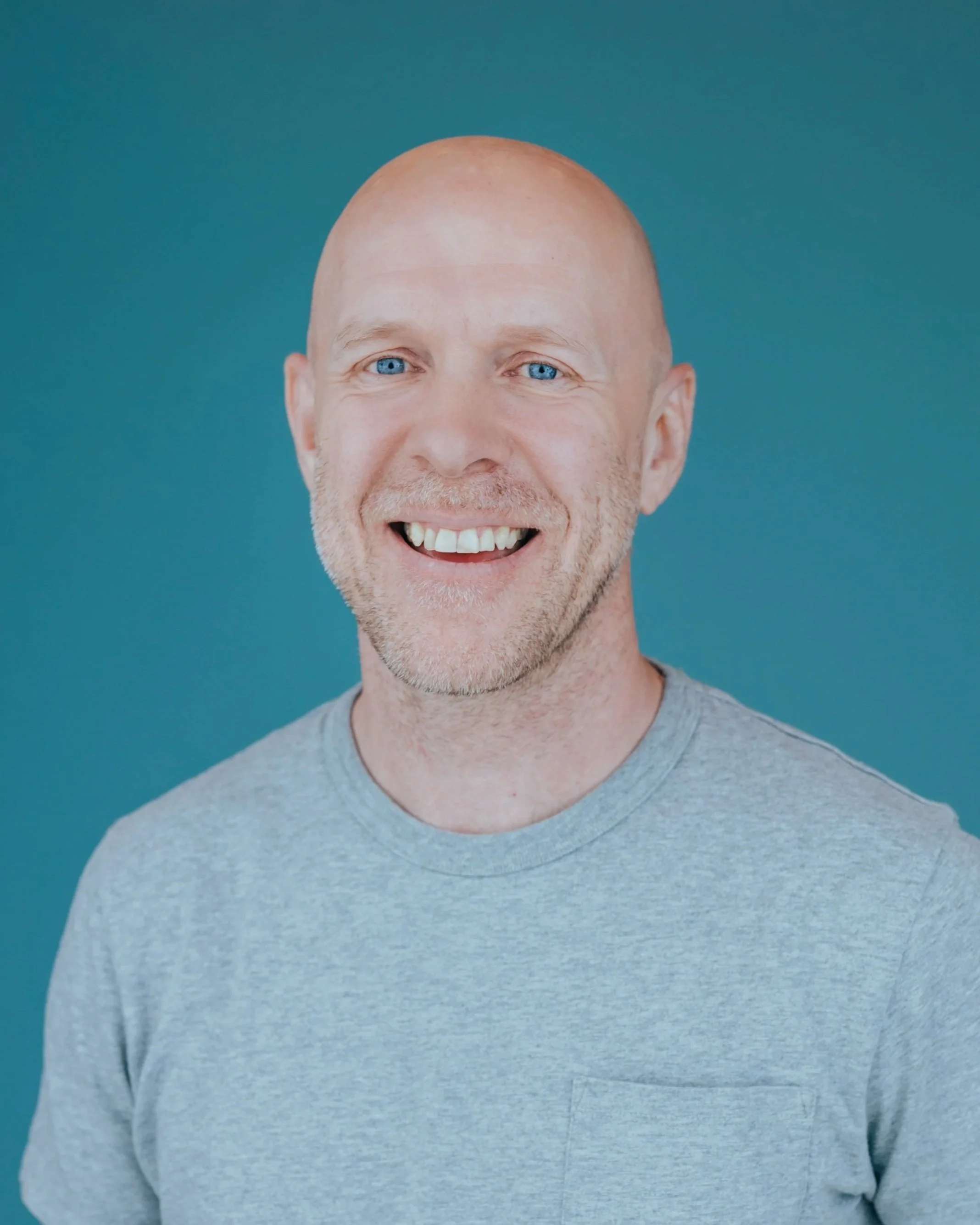 Close-up of a smiling man with blue eyes, bald head, light beard, and wearing a grey t-shirt against a teal background.