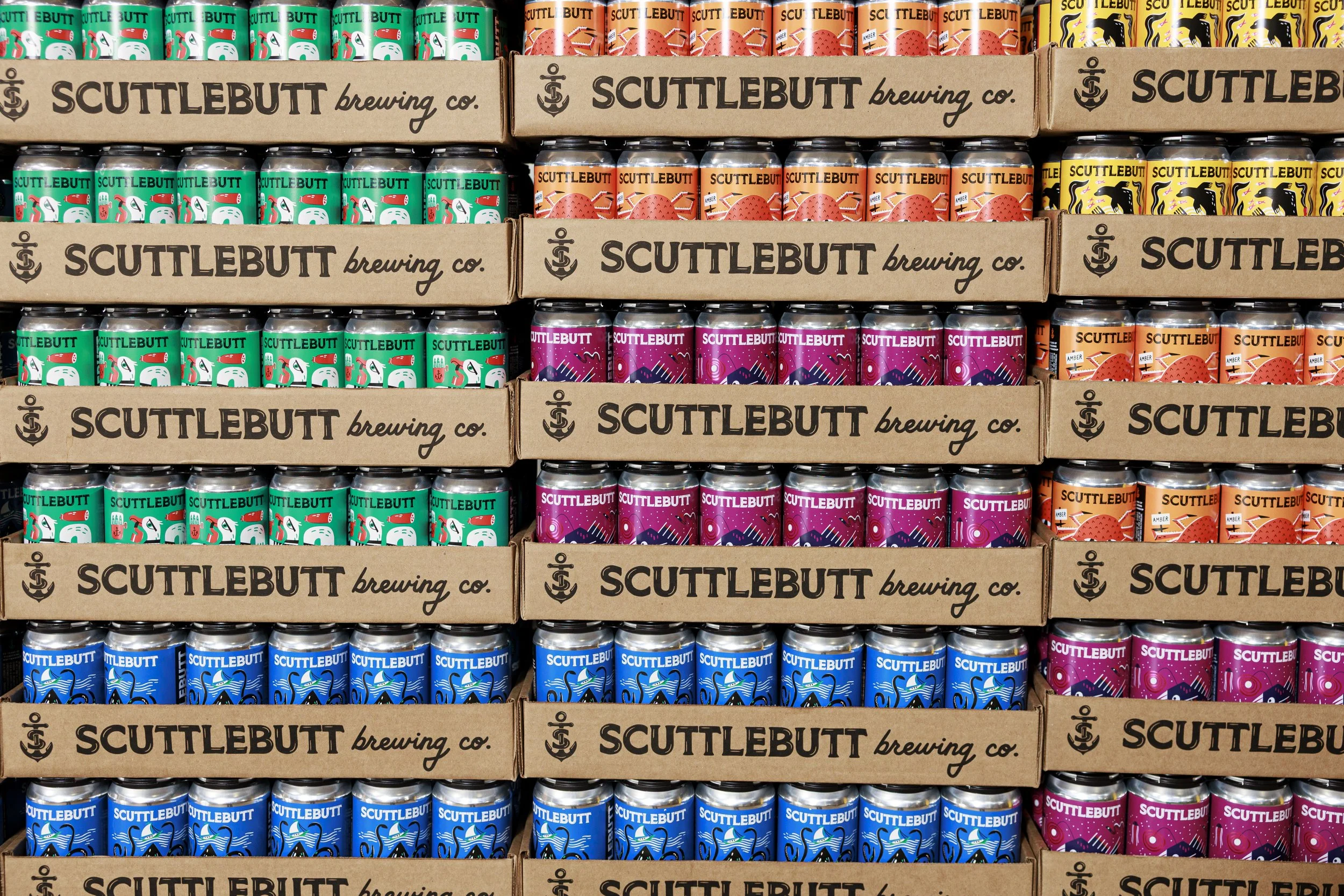 Shelves stocked with various colorful cans of beverage from Scuttlebutt Brewing Co., organized in cardboard boxes with the company's logo and name.