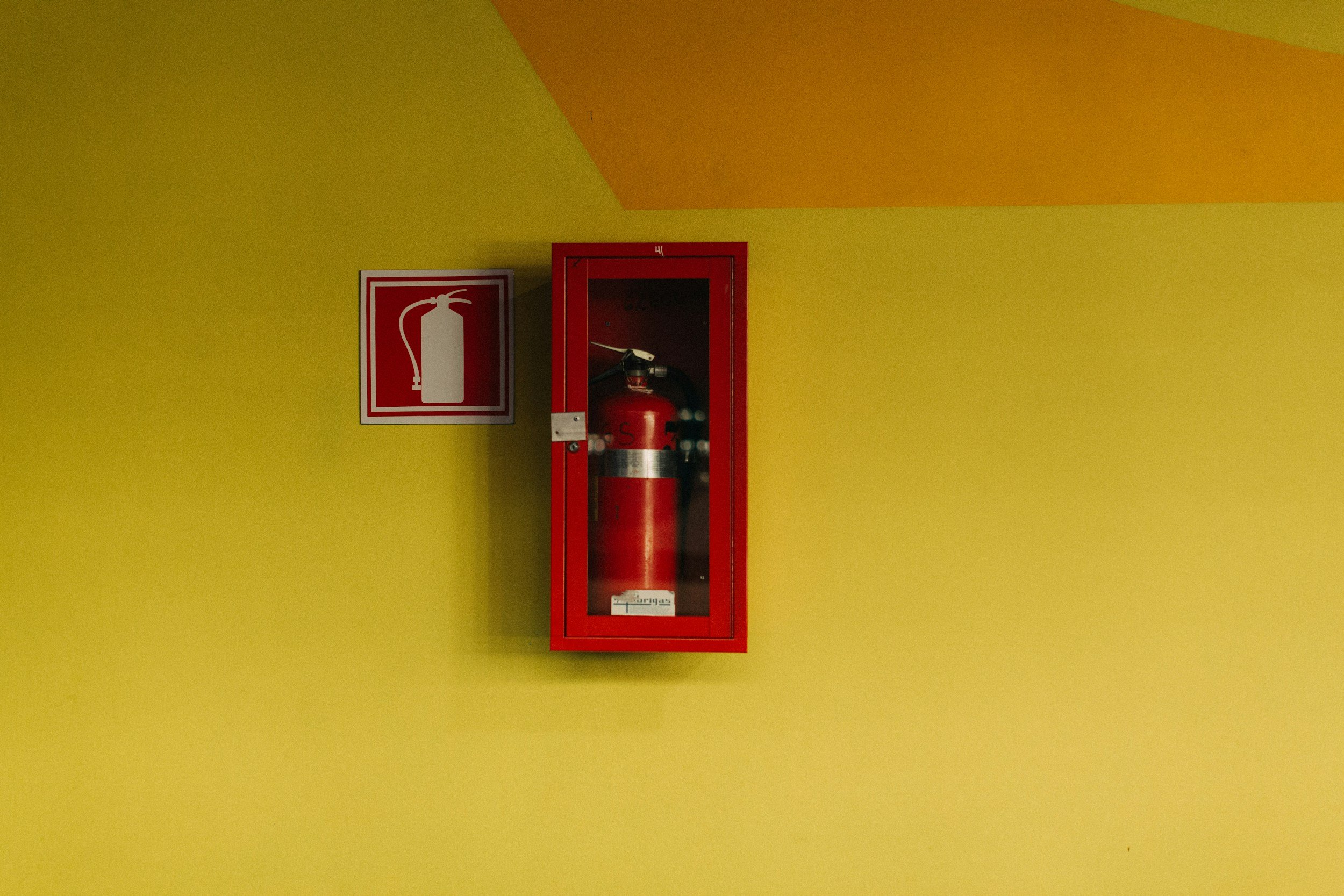 Red fire extinguisher in a wall-mounted cabinet next to a fire extinguisher symbol on a yellow wall.