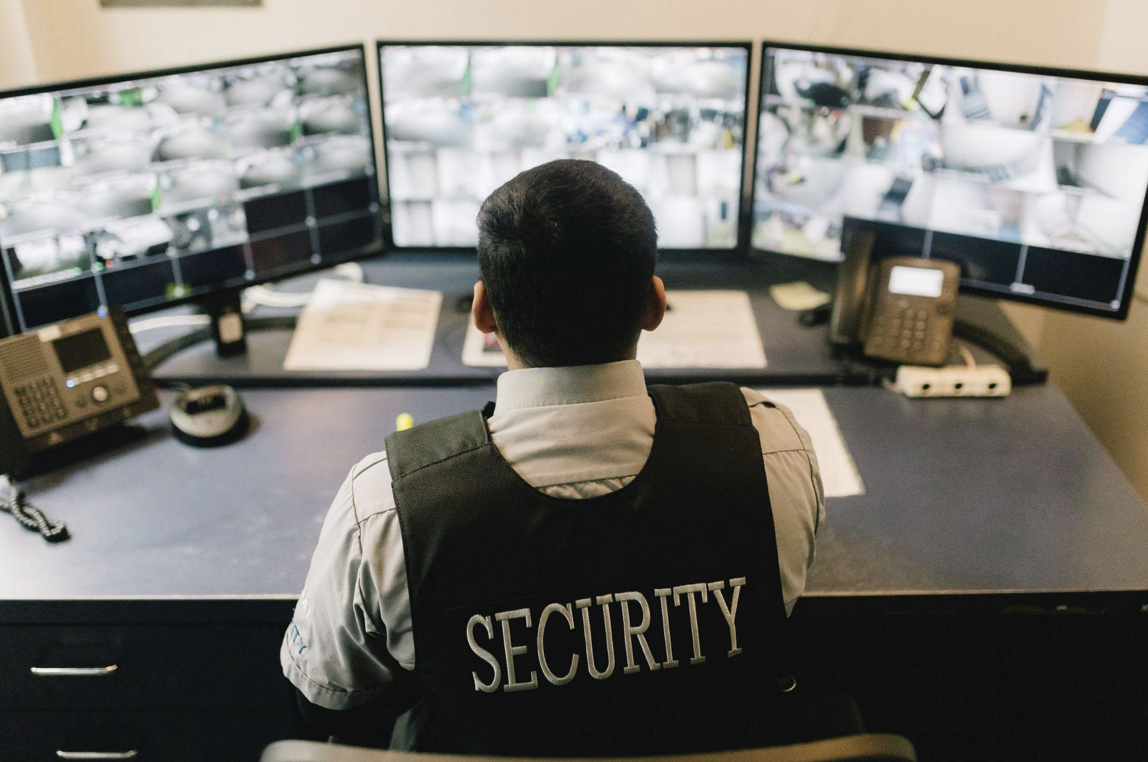 A Regal Security guard sitting at a desk and surveying three monitors displaying multiple security camera feeds.