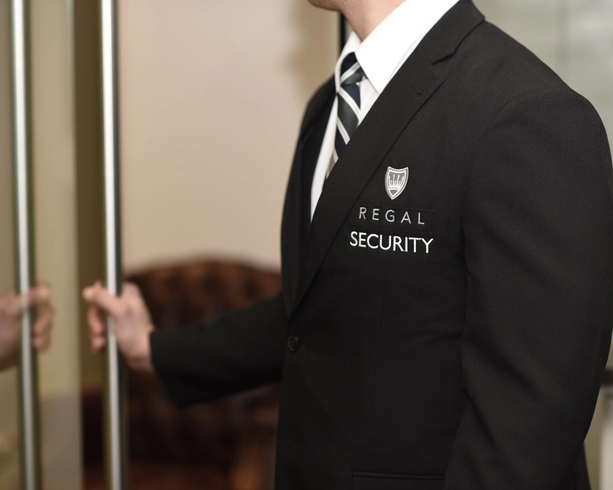Security guard in black suit with "Regal Security" logo, holding a door handle.