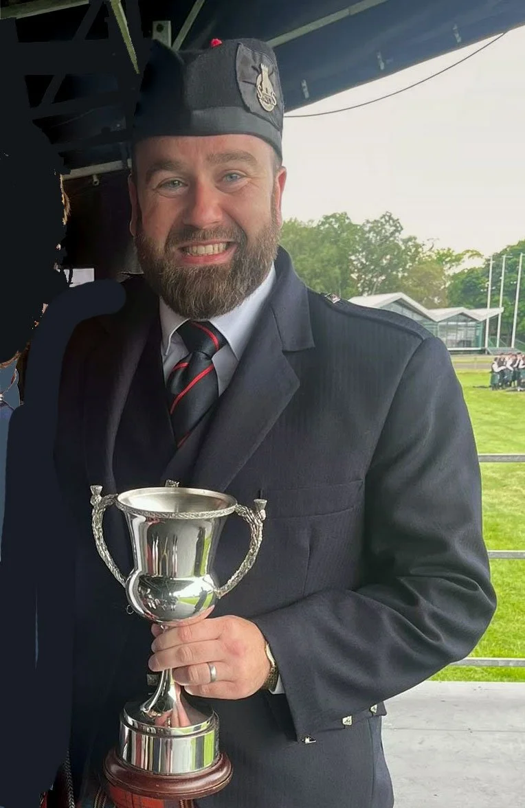 Smiling man in a suit with a bagpipe, outdoors with trees and cloudy sky in the background.