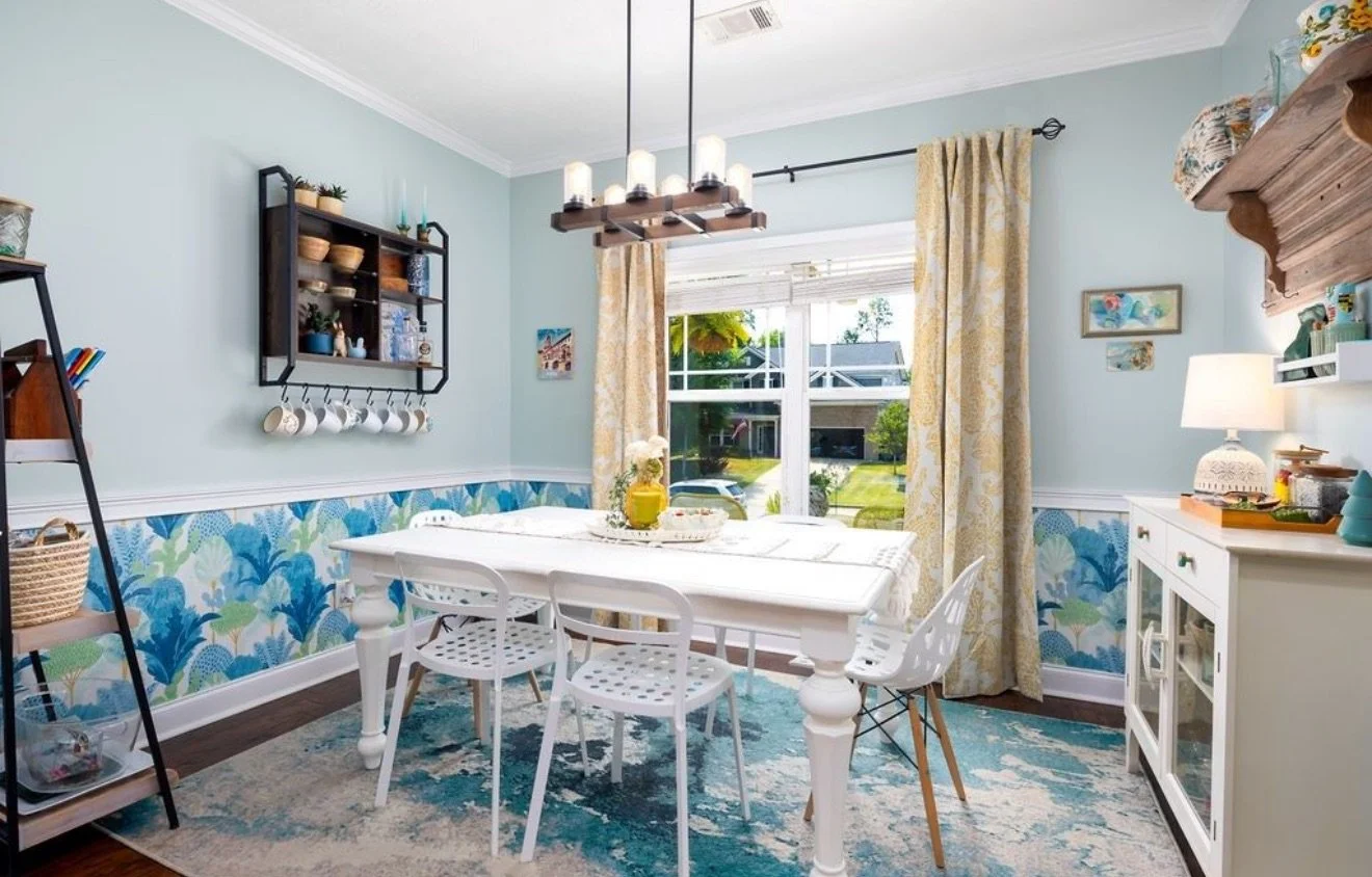 A bright dining room with a white table and mixed white and wooden chairs. There's a window with patterned curtains, a decorative rug, a side cabinet with a table lamp, and wall art. A black wall shelf with cups and bowls is on the wall, and a black ladder shelf with baskets and colorful items is in the corner.