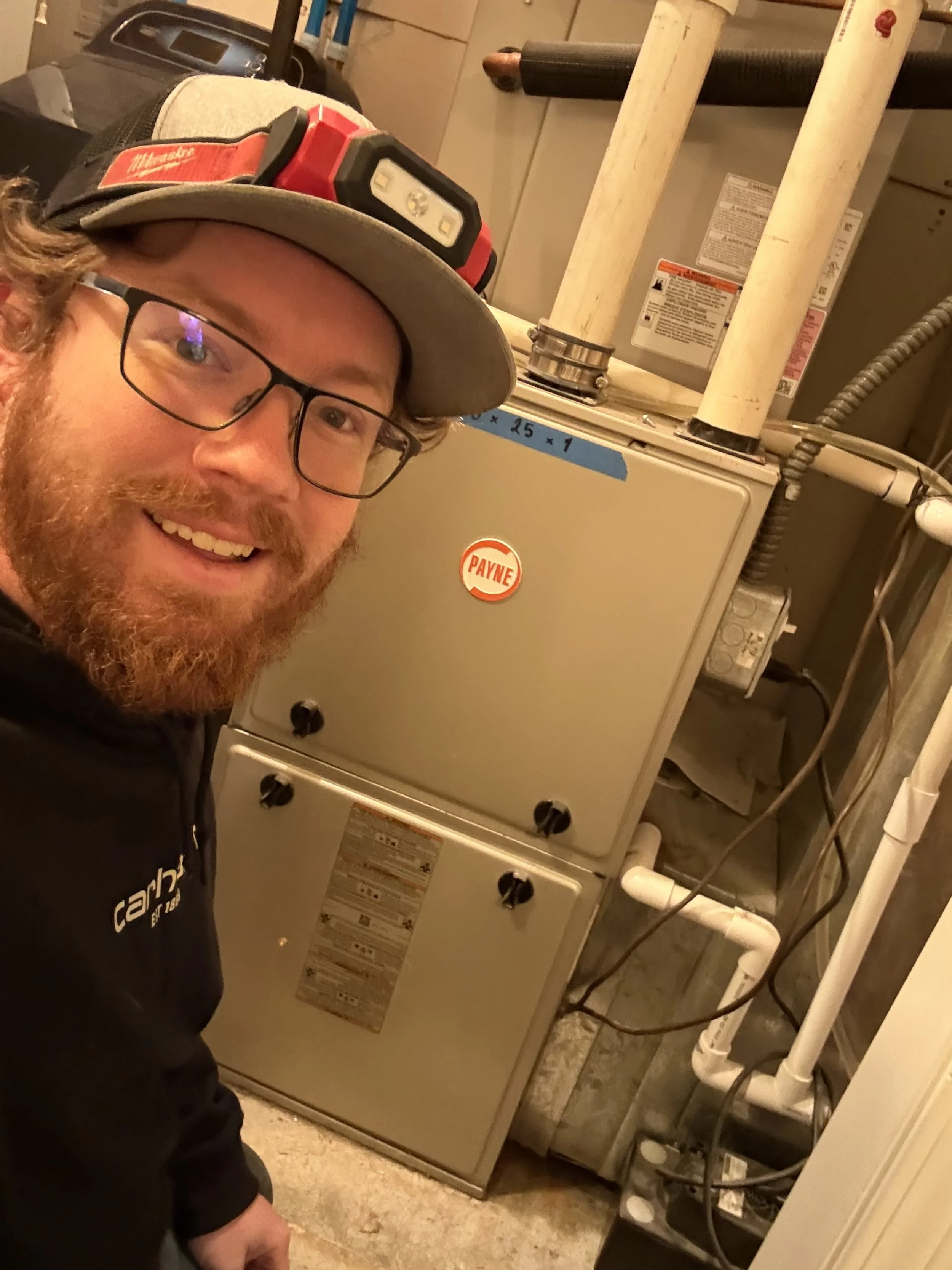 A man with glasses, a beard, and a hard hat with a headlamp, smiling for a selfie in front of HVAC equipment in a basement utility room.